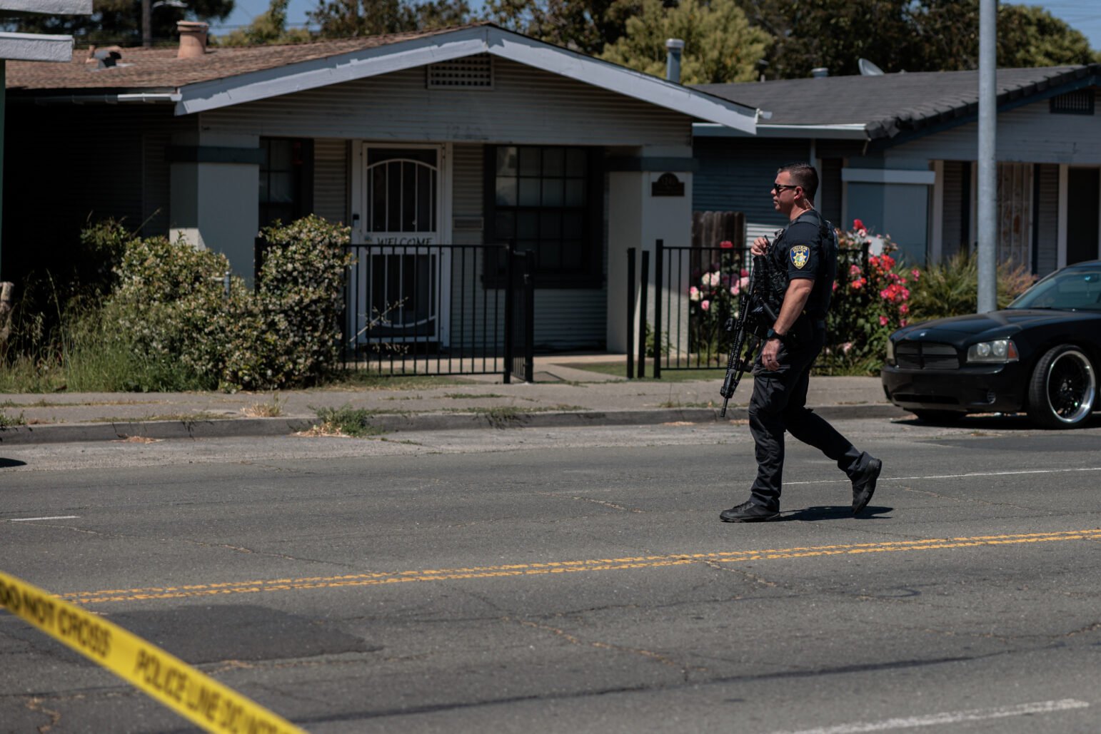 A police officer in tactical vest and sunglasses carries a rifle while crossing a cordoned-off residential street, yellow crime-scene tape in the foreground.