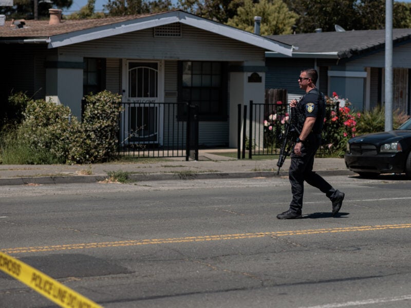 A police officer in tactical vest and sunglasses carries a rifle while crossing a cordoned-off residential street, yellow crime-scene tape in the foreground.