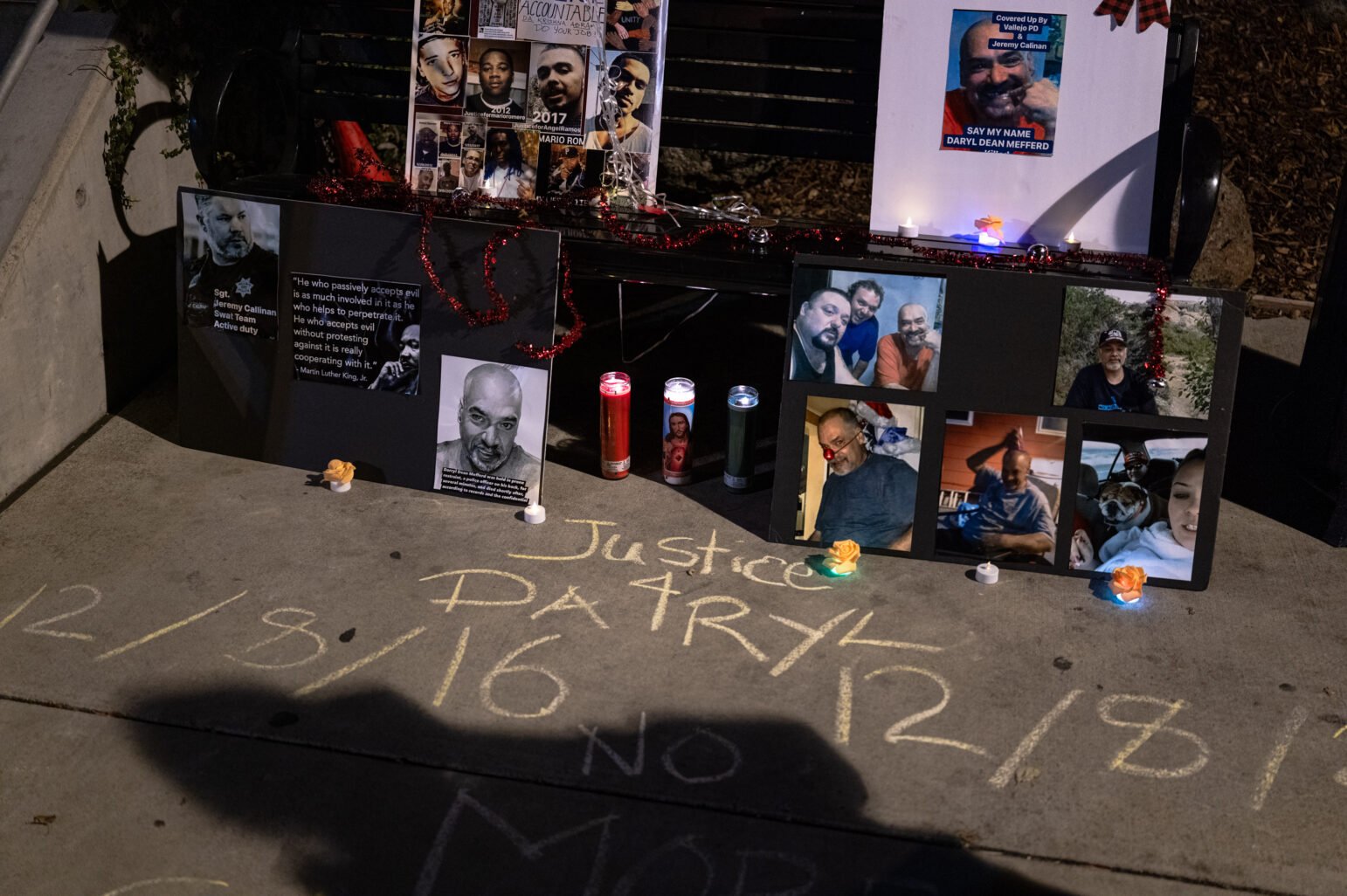 A nighttime sidewalk memorial for Darryl Mefferd: poster boards display photos of Mefferd and others, one naming Vallejo police Sgt. Jeremy Callinan beside a Martin Luther King Jr. quote. Three prayer candles, battery-powered tea lights and small orange paper roses glow in front, while large yellow-chalk letters on the concrete read, “Justice 4 Darryl 12/8/16.”