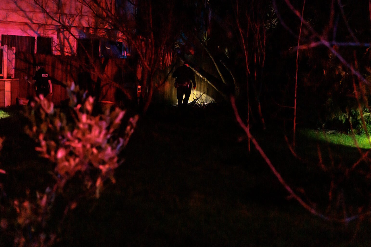 Nighttime image showing Vallejo Police Sgt. Jeremy Callinan walking through a dimly-lit backyard, illuminated partially by police lights and his flashlight. Trees and foliage cast heavy shadows around him.
