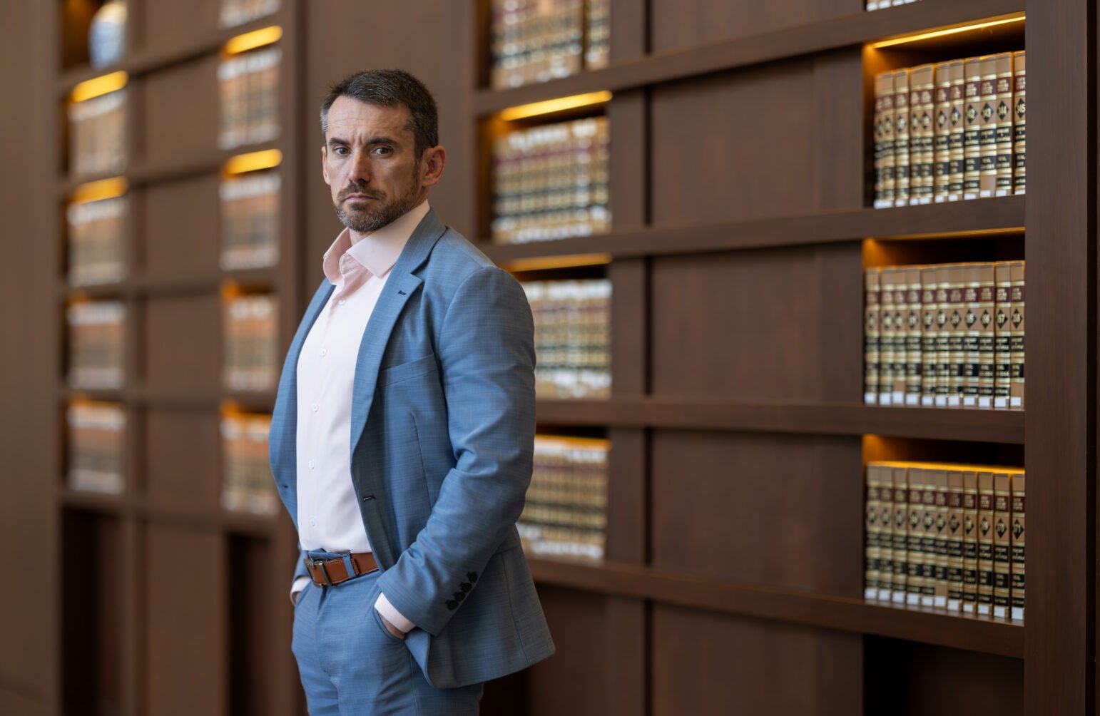 Portrait of Seth Stoughton in a suit, standing confidently in front of shelves filled with legal books. He looks directly into the camera with a serious expression.