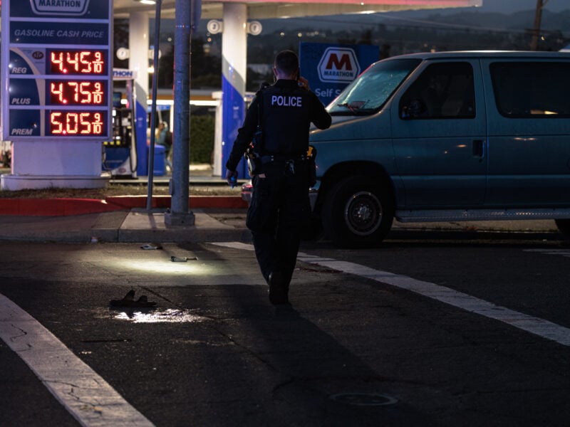 A police officer in a “POLICE” vest walks toward a van at a Marathon gas station at dusk; a lone sandal and small wet puddle lie illuminated on the asphalt in the foreground.