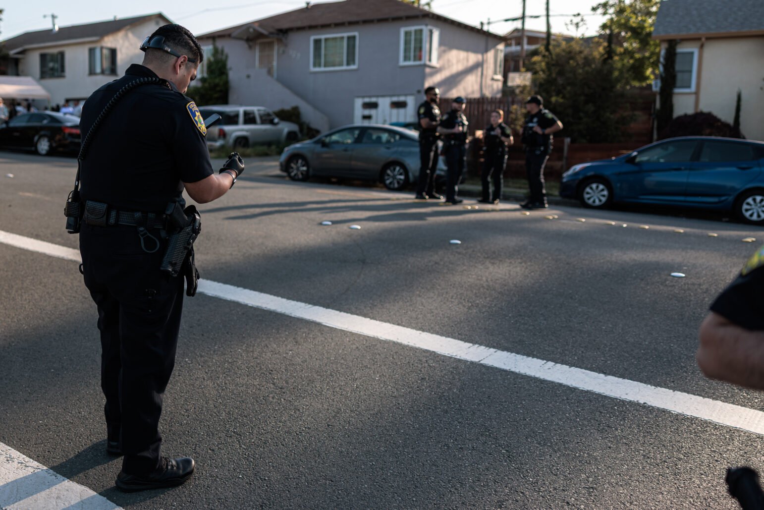 A policeman who wears gloves photographs a used spherical housing in a residential street, while several colleagues are in the background near parked cars.