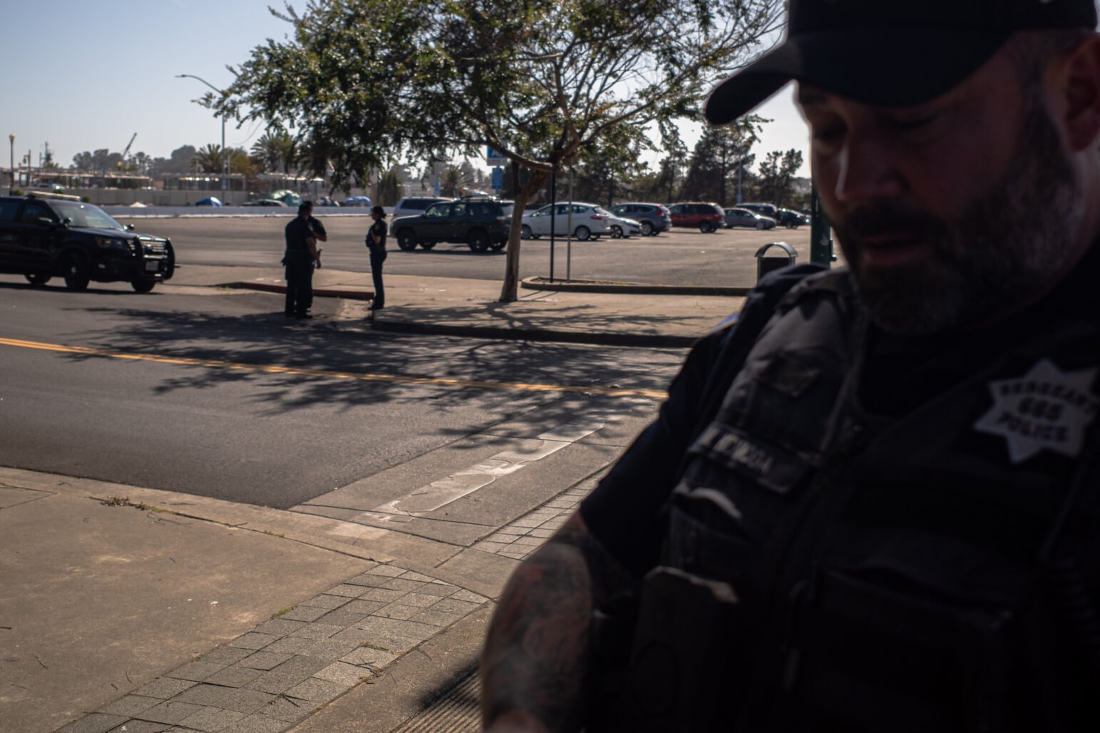 A police officer from Vallejo in Body armor dominates the right foreground, which is somewhat out of focus, while two colleagues are on the water over a sunny road in the city center near a parking space. A black patrol -SUV is parked next to them.