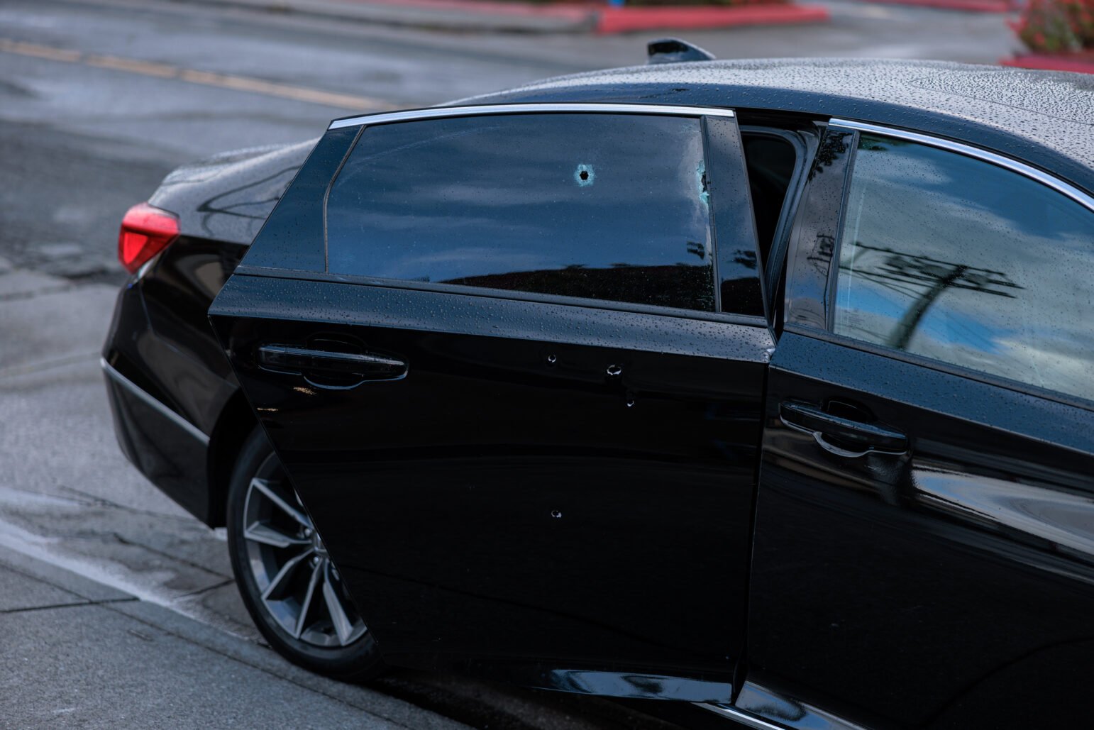 A black sedan with its rear driver-side door ajar shows multiple bullet holes in the door and window. The damage appears fresh, suggesting a recent shooting.