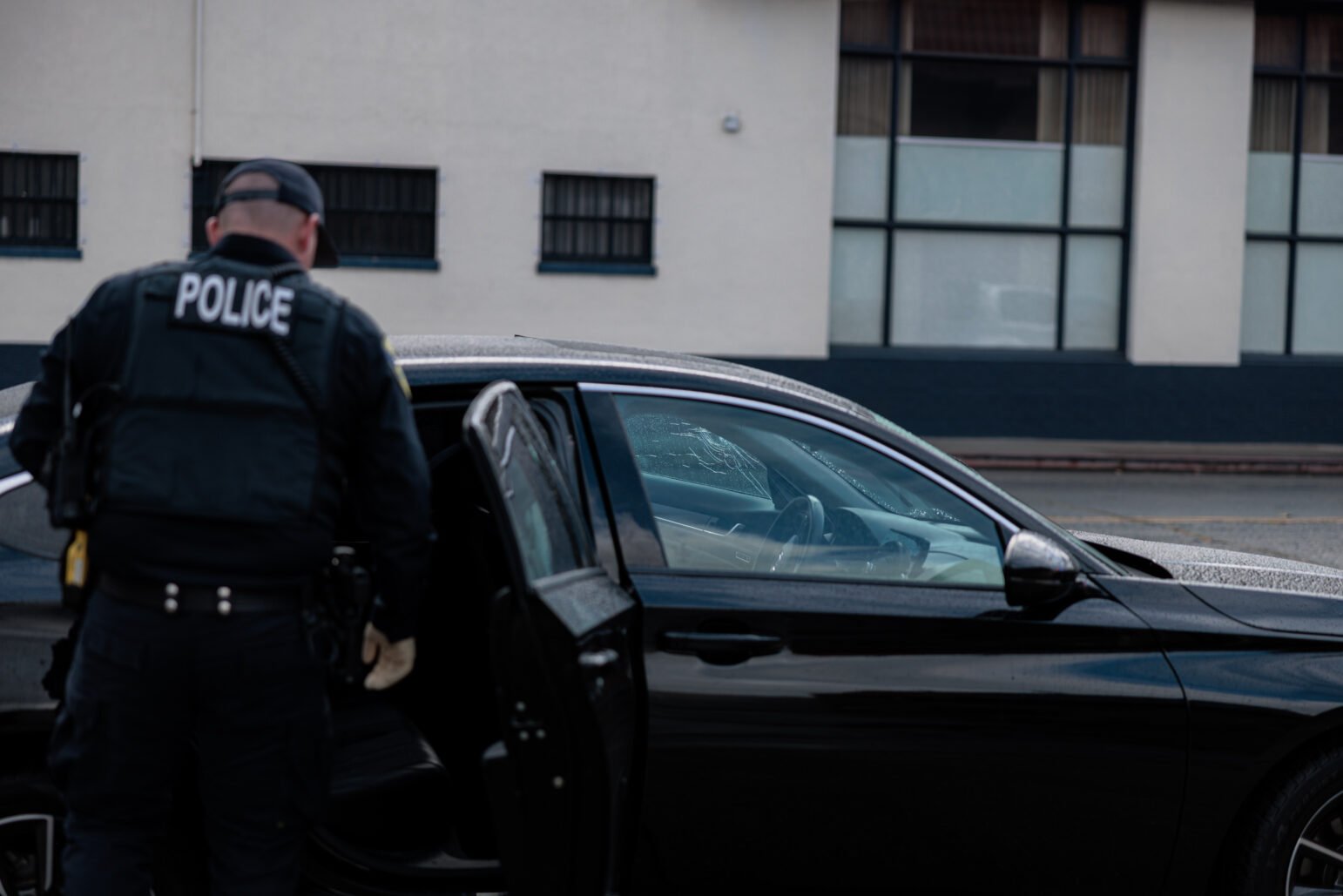 A Vallejo police officer in tactical gear stands beside an open car door, examining the interior of a black sedan. The front passenger window is shattered, with visible cracks and holes in the glass.