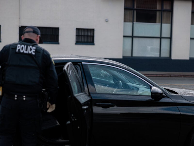 A Vallejo police officer in tactical gear stands beside an open car door, examining the interior of a black sedan. The front passenger window is shattered, with visible cracks and holes in the glass.