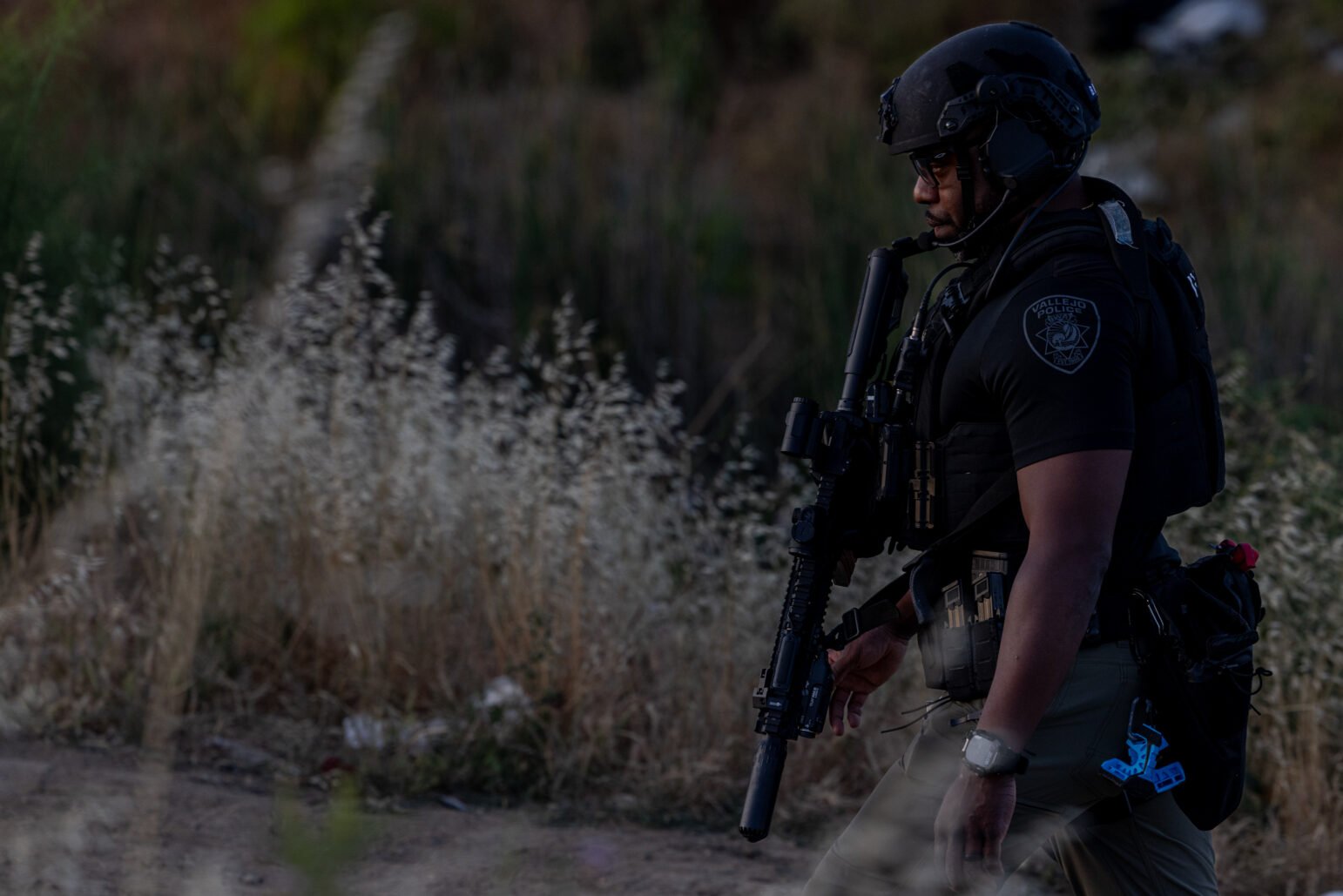 A Vallejo police officer in tactical gear walks through a grassy field at dusk, carrying a rifle and wearing a ballistic helmet and body armor. The officer’s uniform patch is visible, and the surroundings suggest an ongoing emergency response.