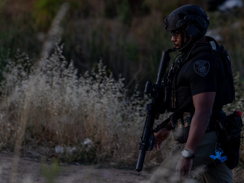 A Vallejo police officer in tactical gear walks through a grassy field at dusk, carrying a rifle and wearing a ballistic helmet and body armor. The officer’s uniform patch is visible, and the surroundings suggest an ongoing emergency response.