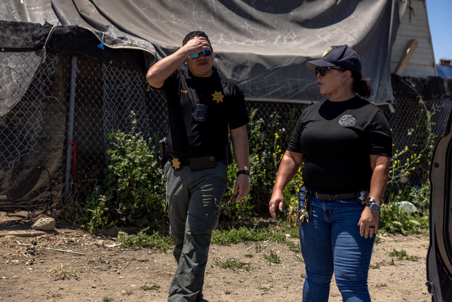 Two law enforcement officials walk past a makeshift structure covered in tarps and fencing. The male official shields his eyes from the sun while the female officer speaks.