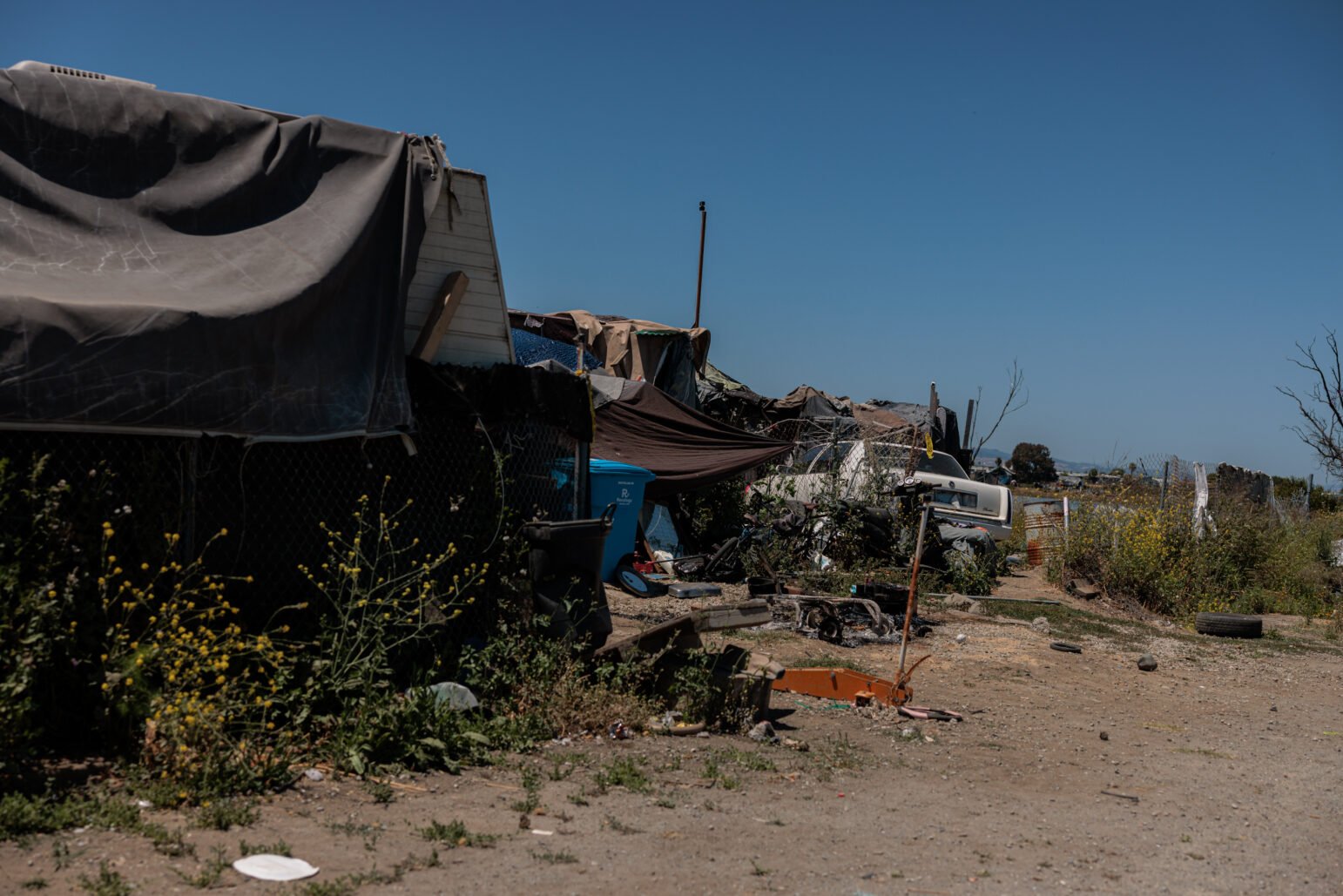 An encampment constructed from tarps, fencing, and scrap materials stretches along a dirt path under a bright blue sky. Debris and vegetation are scattered throughout the area, including an overturned scooter and abandoned appliances.
