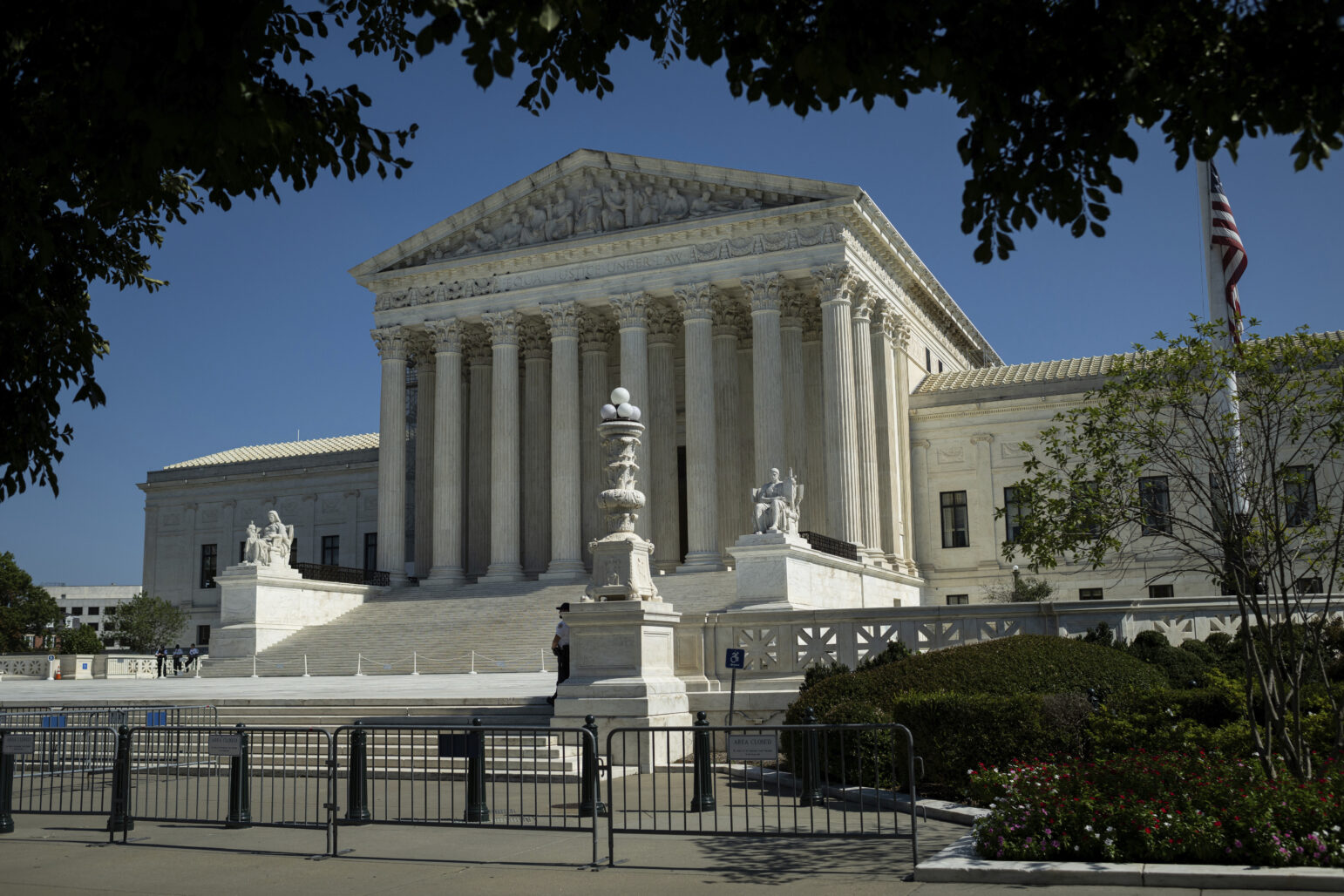 A wide view of the U.S. Supreme Court building in Washington, D.C., showing its iconic white marble facade, tall Corinthian columns, and broad steps leading to the entrance. Statues flank the staircase, and temporary barriers are placed along the sidewalk in front. An American flag stands to the right, and tree branches frame the image at the top.