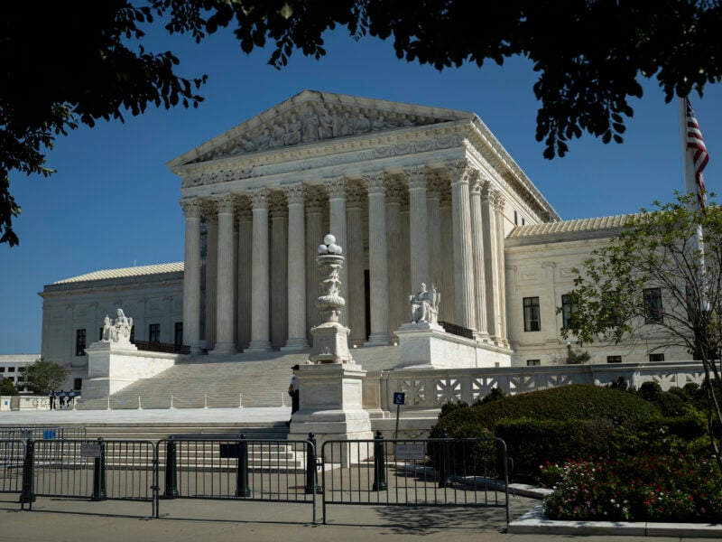 A wide view of the U.S. Supreme Court building in Washington, D.C., showing its iconic white marble facade, tall Corinthian columns, and broad steps leading to the entrance. Statues flank the staircase, and temporary barriers are placed along the sidewalk in front. An American flag stands to the right, and tree branches frame the image at the top.