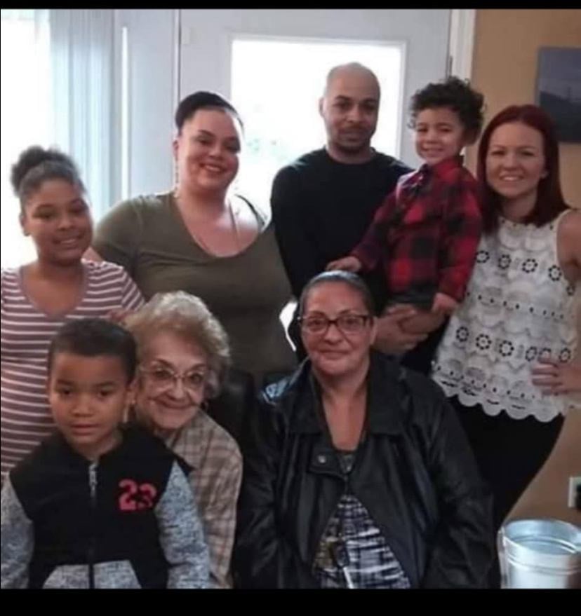 A group of eight people from multiple generations pose in a warmly lit home. Evangelina R. Humphrey is seated near the center, surrounded by children, adults, and a toddler in a red plaid shirt held by an adult.