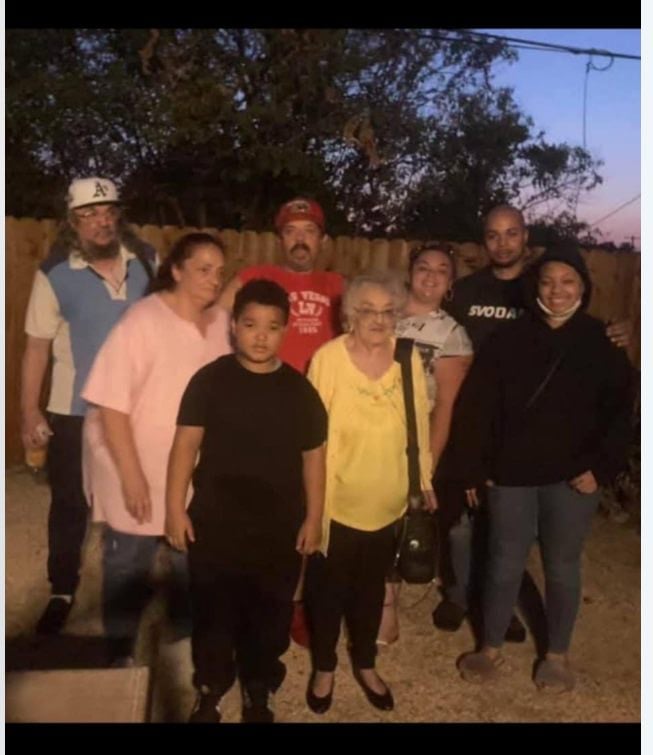 A group of eight family members poses for a photo in a backyard at dusk. An elderly woman in a yellow top stands at the center, surrounded by adults and children of various ages, all standing in front of a wooden fence.