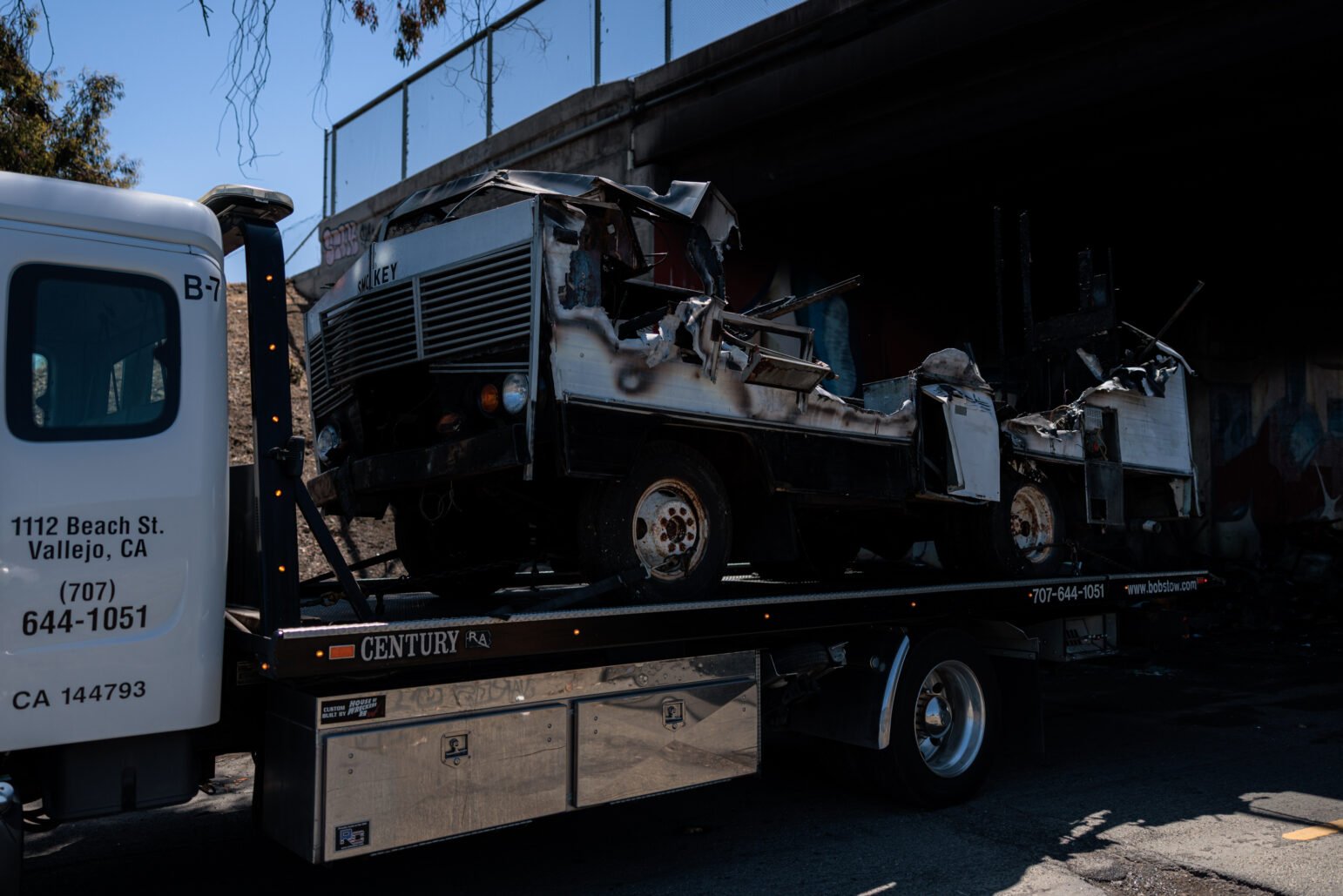 A severely burned motor home is loaded onto a flatbed tow truck beneath an overpass in Vallejo, California. The vehicle is extensively damaged, with its roof and sides partially melted and collapsed. The word “Smokey” is visible on the front grille. The tow truck displays a local company’s name and contact information.
