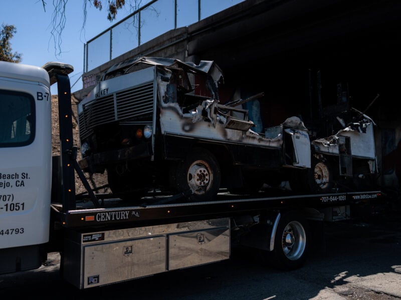 A severely burned motor home is loaded onto a flatbed tow truck beneath an overpass in Vallejo, California. The vehicle is extensively damaged, with its roof and sides partially melted and collapsed. The word “Smokey” is visible on the front grille. The tow truck displays a local company’s name and contact information.