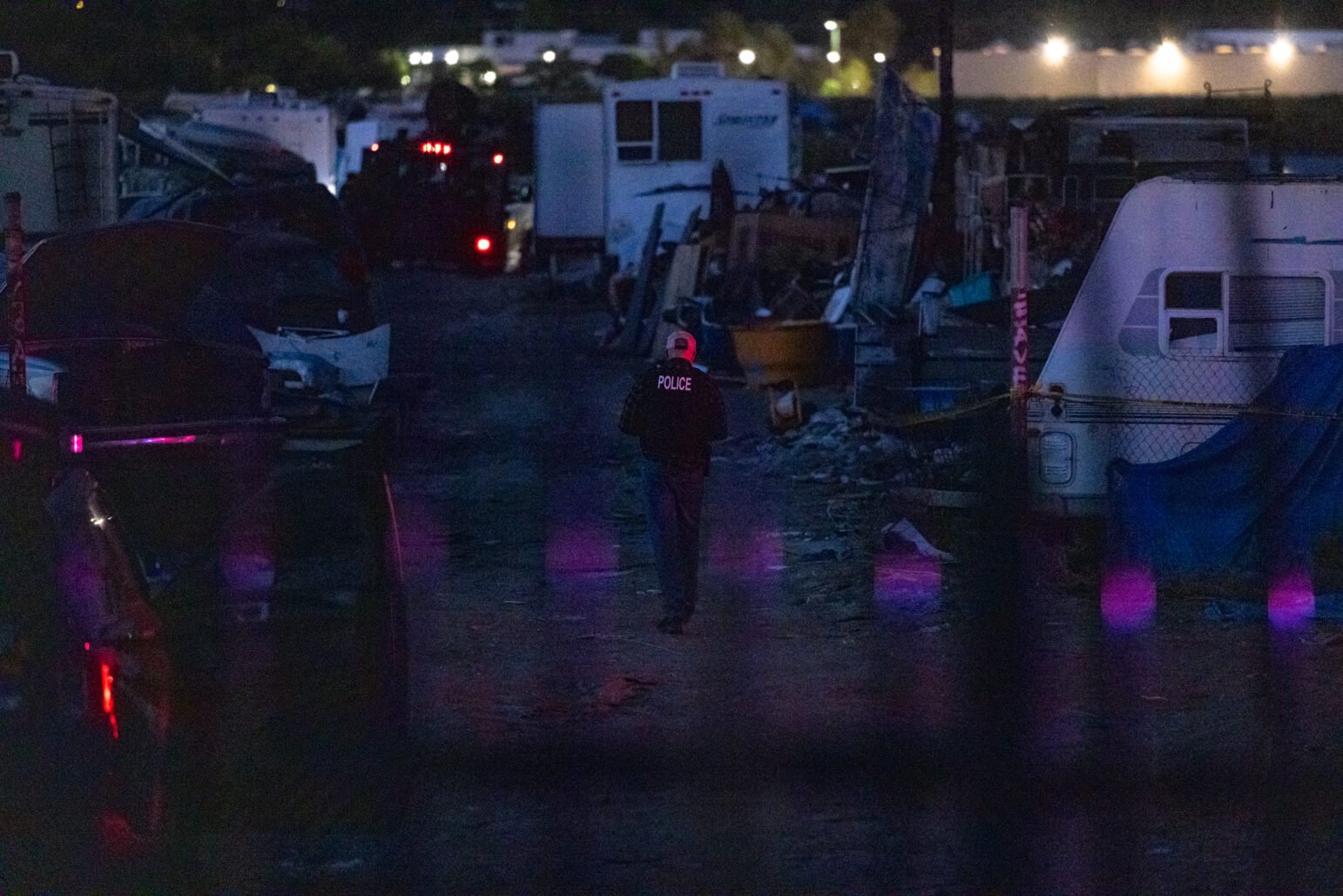 A police officer wearing a jacket labeled “POLICE” walks alone through a cluttered encampment at night, surrounded by trailers, debris, and abandoned vehicles, illuminated by dim ambient light and flashes of red and blue from emergency lights.