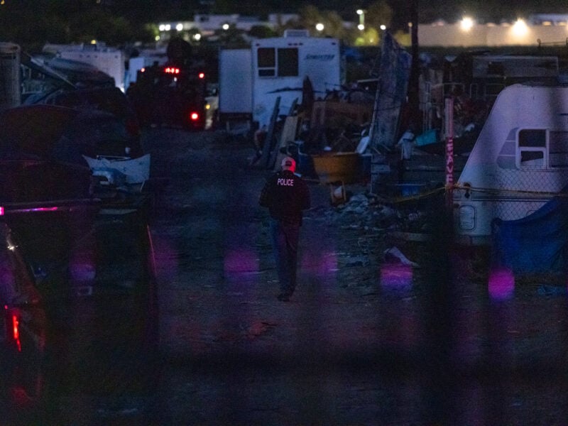 A police officer wearing a jacket labeled “POLICE” walks alone through a cluttered encampment at night, surrounded by trailers, debris, and abandoned vehicles, illuminated by dim ambient light and flashes of red and blue from emergency lights.