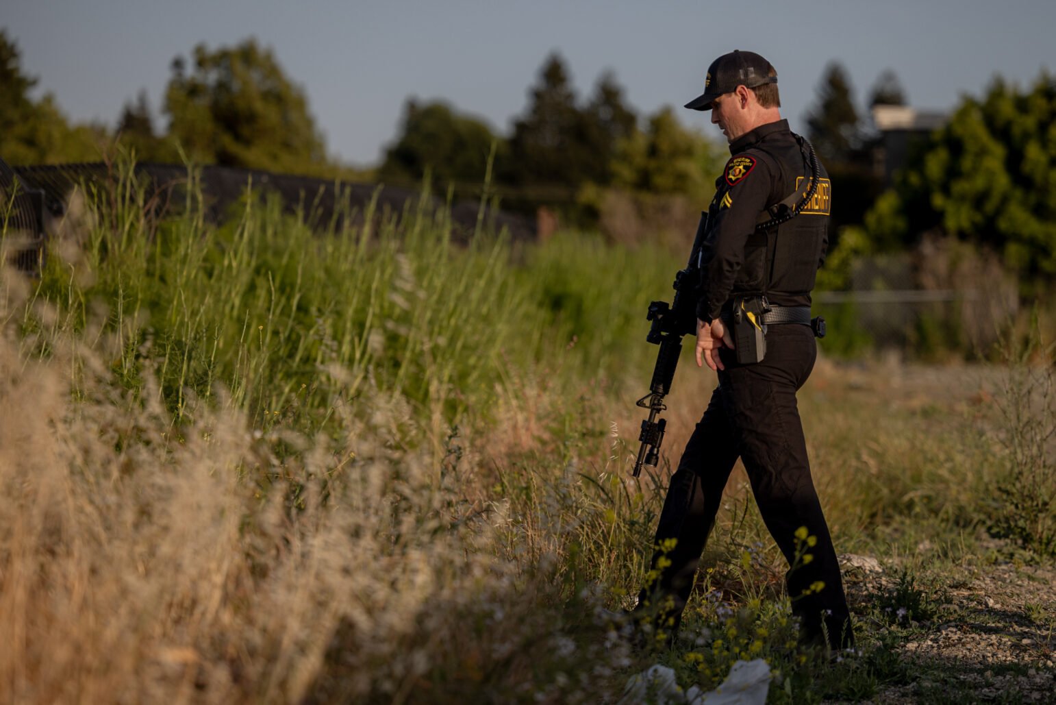 A uniformed sheriff’s deputy holding a rifle stands in tall grass, observing an area during daylight. He appears focused and attentive.