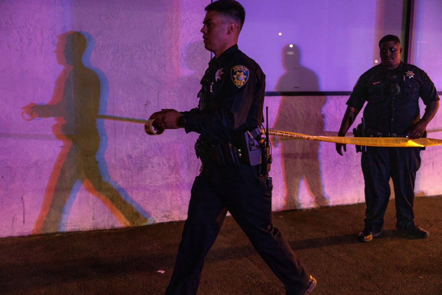 A Vallejo police officer unspools yellow crime scene tape outside a building at night, illuminated by flashing red and blue emergency lights. Another officer stands nearby against a wall, casting overlapping multicolored shadows behind them.