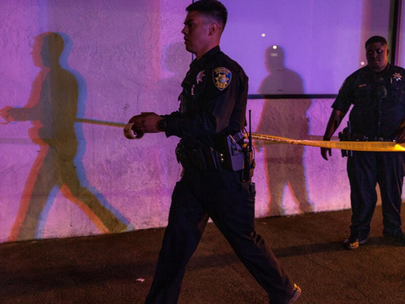 A Vallejo police officer unspools yellow crime scene tape outside a building at night, illuminated by flashing red and blue emergency lights. Another officer stands nearby against a wall, casting overlapping multicolored shadows behind them.