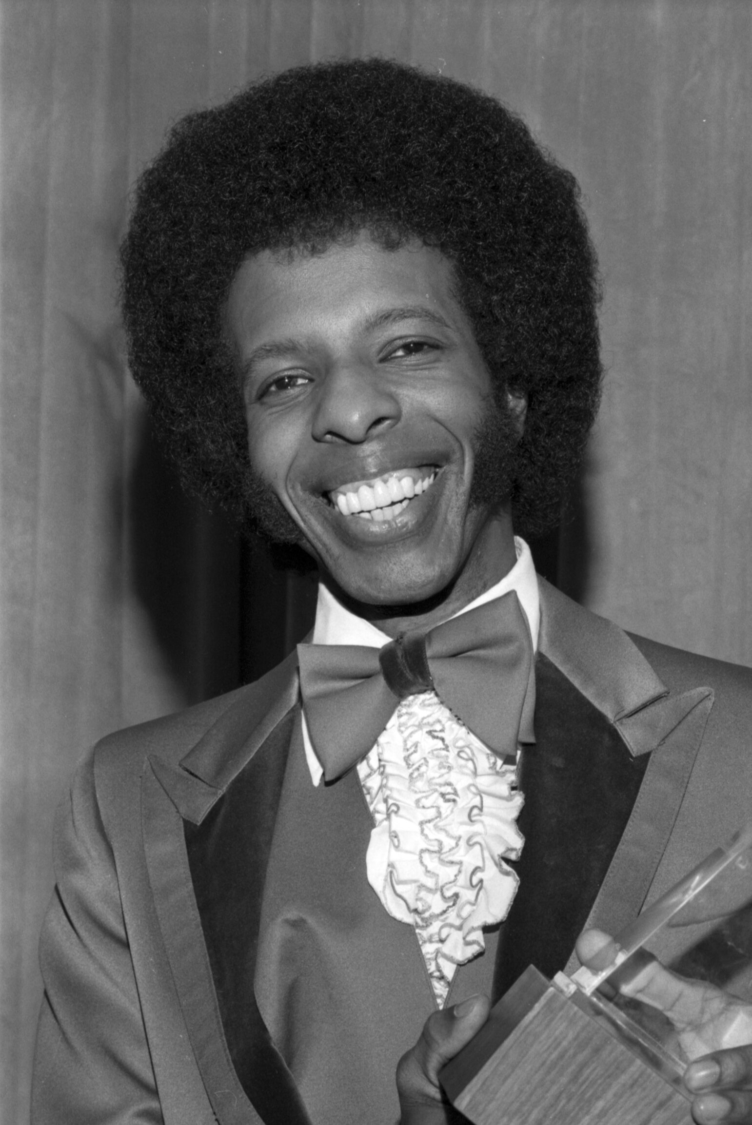 A smiling man dressed in a vintage tuxedo with wide lapels, a bow tie, and a ruffled shirt holds an award. The black-and-white portrait captures him mid-laugh, framed against a plain backdrop.