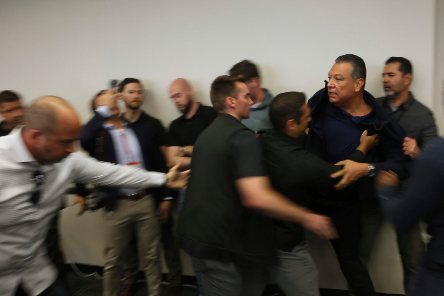 A group of men, some appearing to be plainclothes officers or security personnel, forcibly detain a man against a wall during a chaotic moment indoors. One man grabs the front of the detainee’s shirt while others reach toward him, and several onlookers react or record the scene with phones. The image is slightly blurred, conveying urgency and motion.