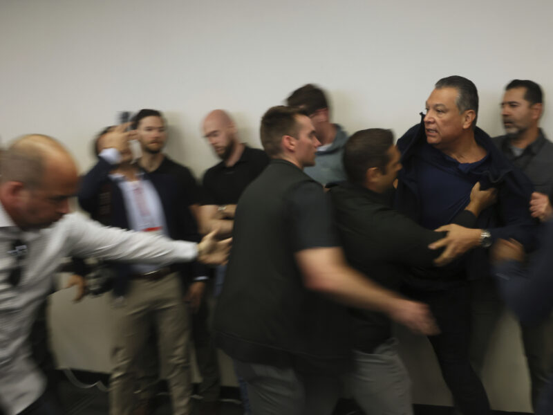 A group of men, some appearing to be plainclothes officers or security personnel, forcibly detain a man against a wall during a chaotic moment indoors. One man grabs the front of the detainee’s shirt while others reach toward him, and several onlookers react or record the scene with phones. The image is slightly blurred, conveying urgency and motion.