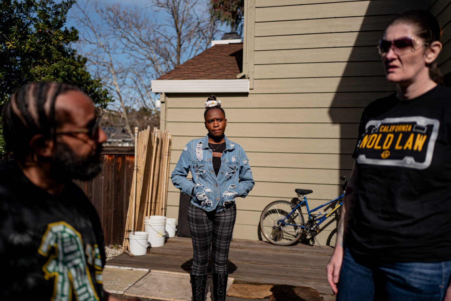 Deyana Jenkins stands with her hands in her pockets in the center of a sunlit yard, flanked by two other individuals. She wears a distressed denim jacket and plaid pants. A bicycle rests against a beige house behind her.