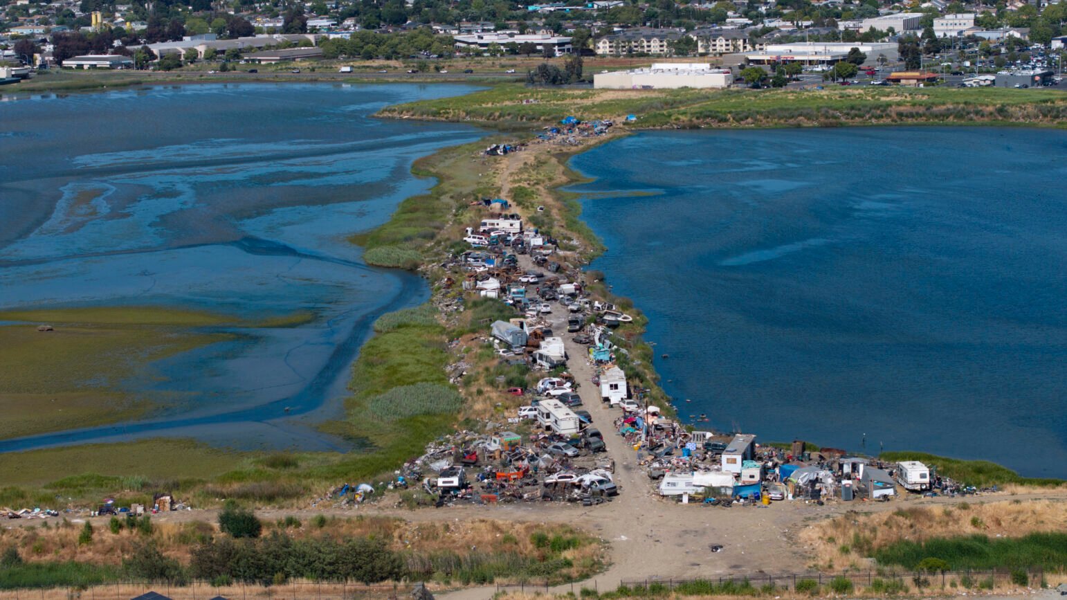 An aerial view shows a narrow strip of land densely packed with vehicles, debris, and makeshift shelters, extending into a body of water surrounded by grassy wetlands and urban areas in the distance.