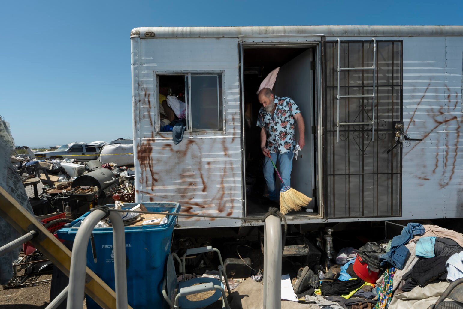 A man wearing a floral-patterned shirt and jeans sweeps debris from the doorway of a metal trailer surrounded by piles of belongings and garbage, with a blue recycling bin nearby.