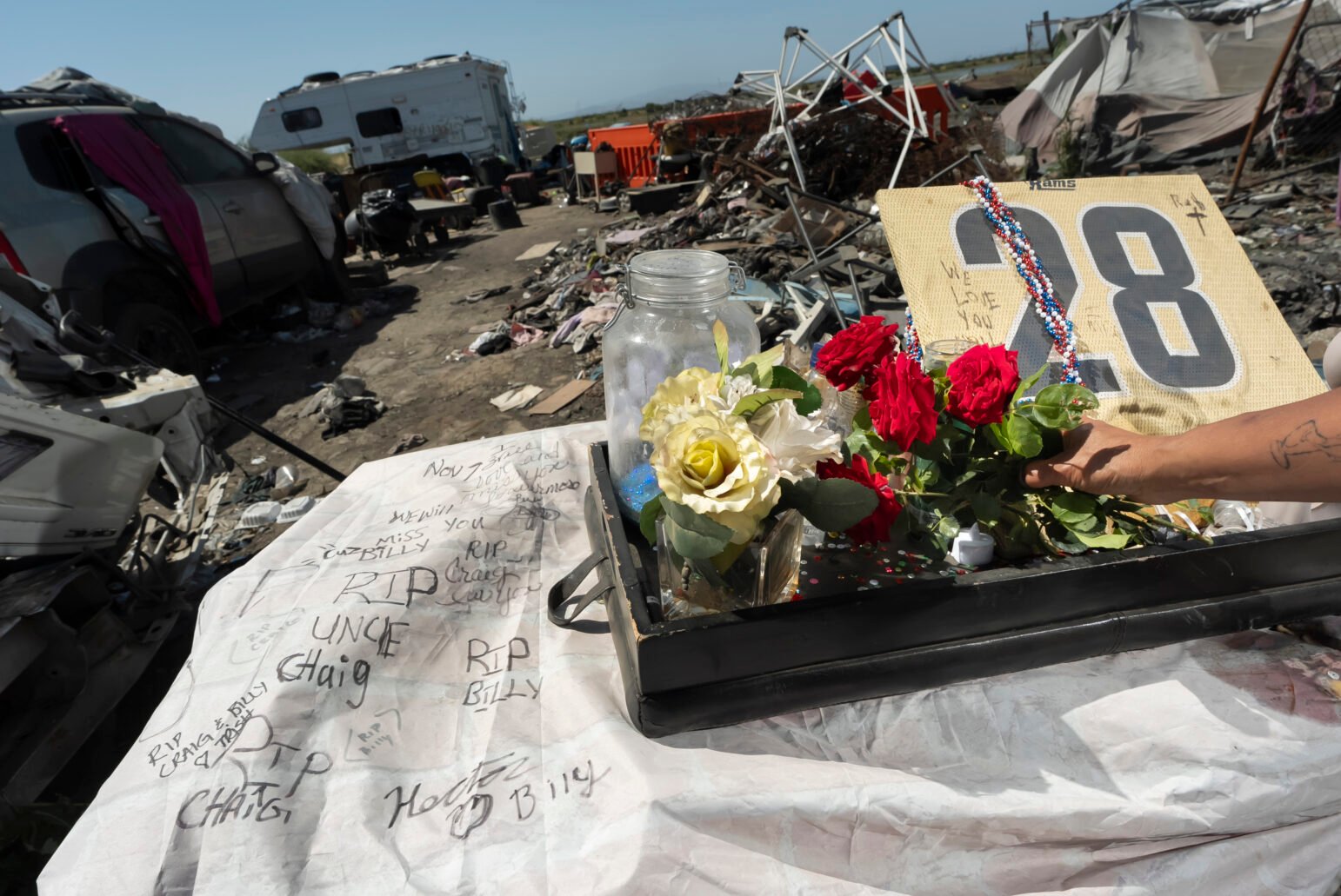 A memorial set on a table amid debris at an encampment. Flowers, candles, a decorated jersey bearing the number 28, and handwritten messages of remembrance are visible.