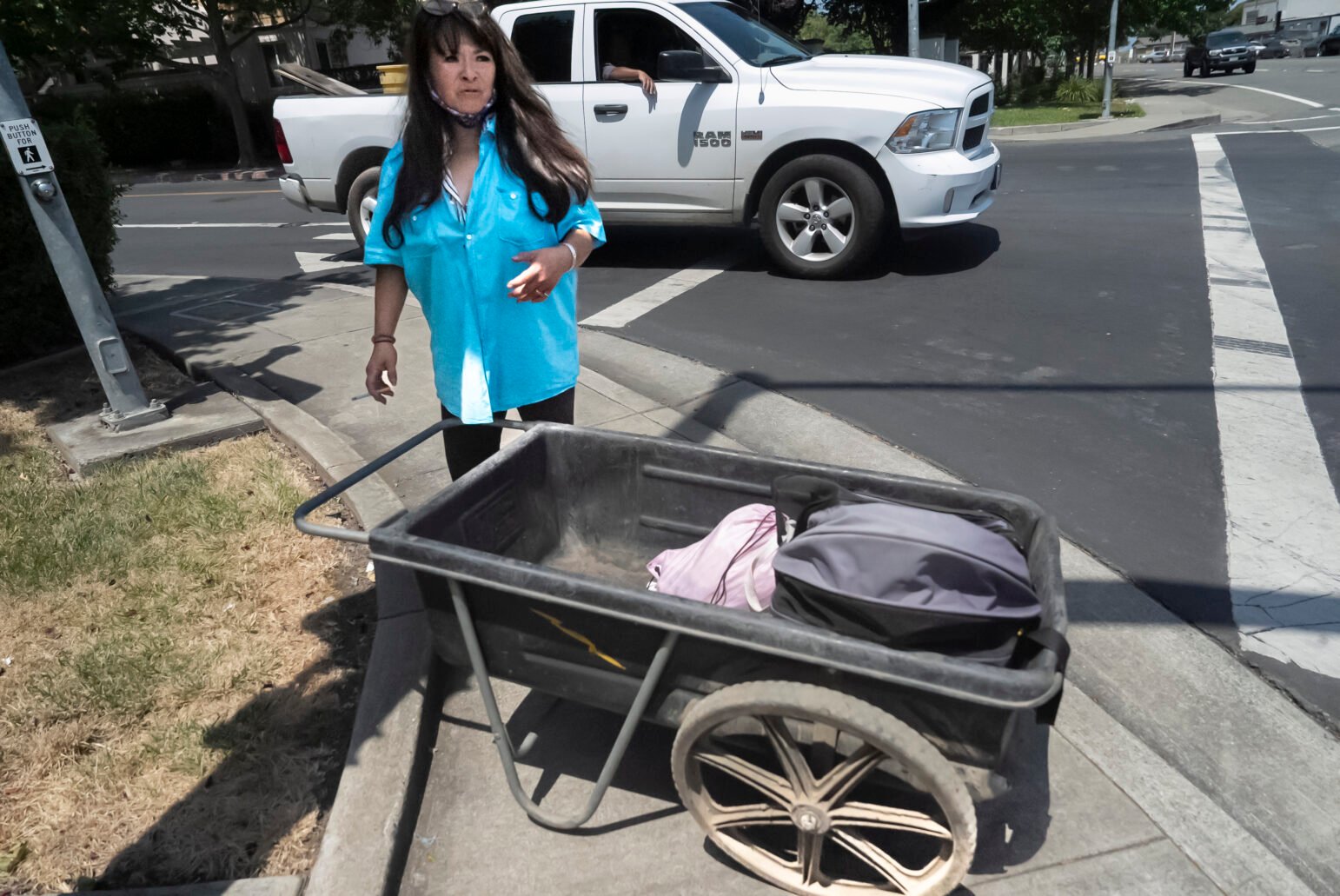 A woman wearing a bright blue shirt stands beside a plastic cart containing personal items at a street corner intersection. A white truck is seen in the background.