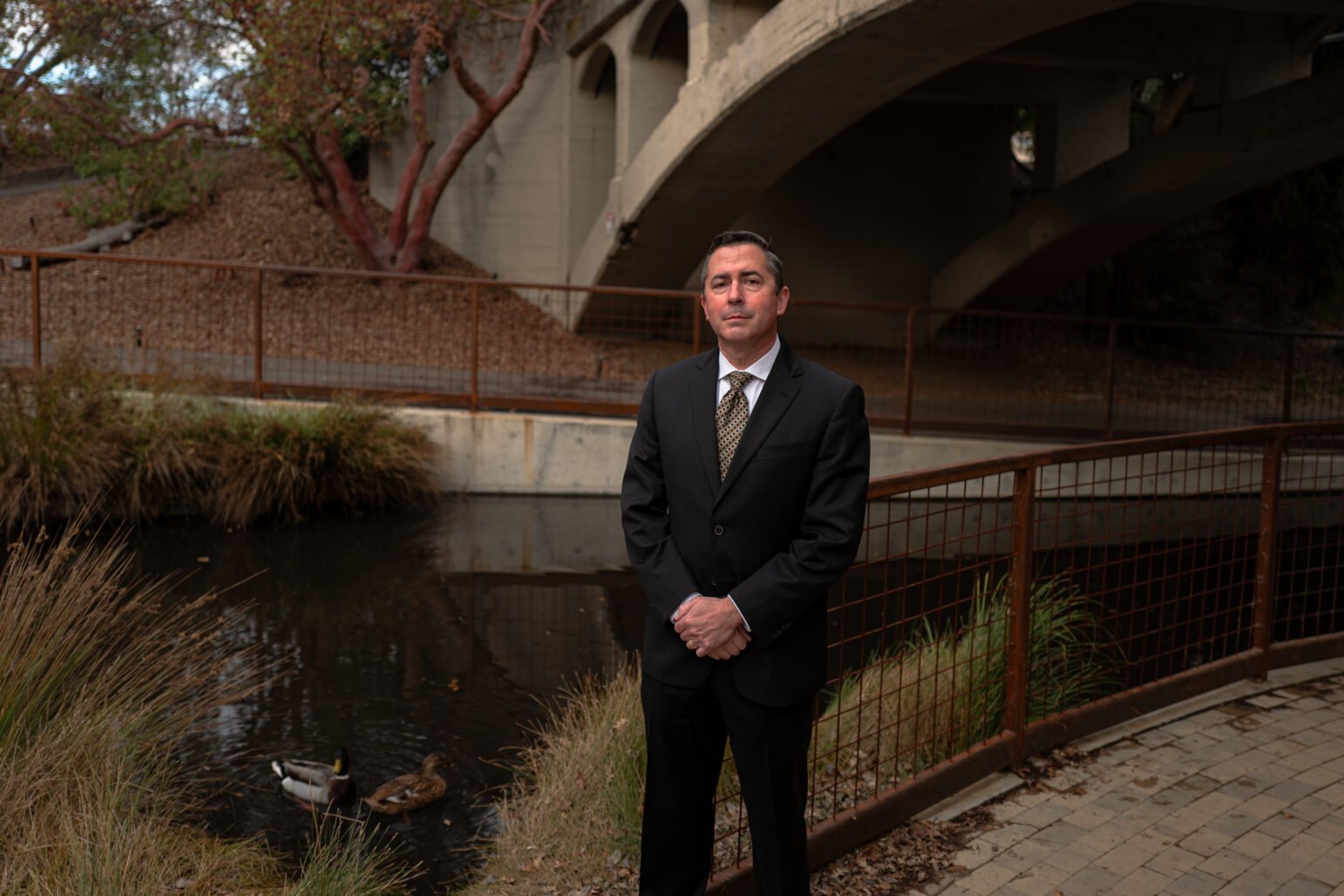 Former Vallejo Police Capt. John Whitney stands in a black suit and tie near a shallow creek, flanked by reeds and ducks, with a concrete bridge arching in the background.