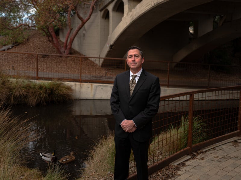 Former Vallejo Police Capt. John Whitney stands in a black suit and tie near a shallow creek, flanked by reeds and ducks, with a concrete bridge arching in the background.