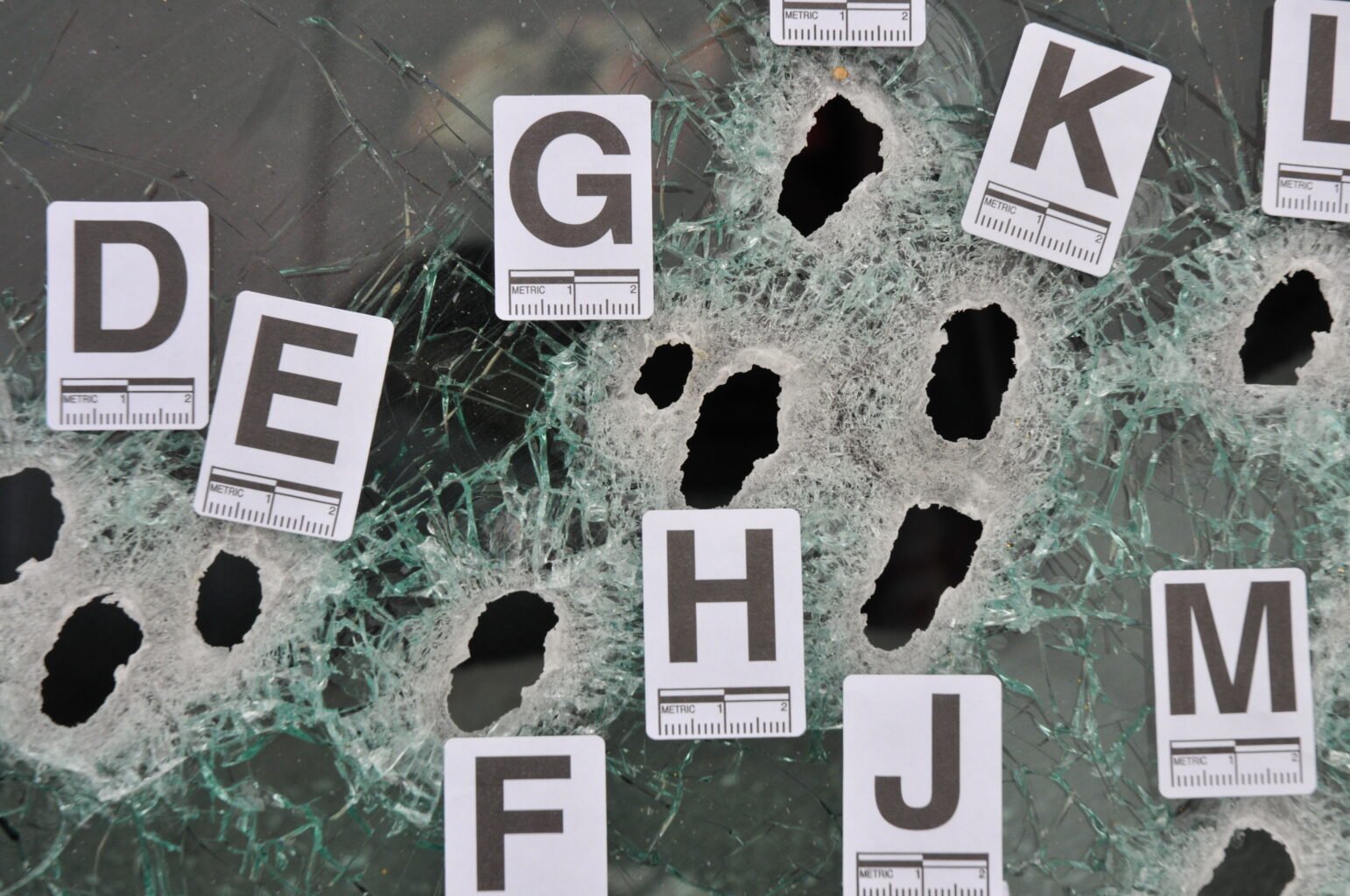 A shattered pane of glass with multiple bullet holes, each labeled with a lettered forensic marker ranging from D to M. Cracks radiate from the impact points, and metric scales are printed on each tag for reference.
