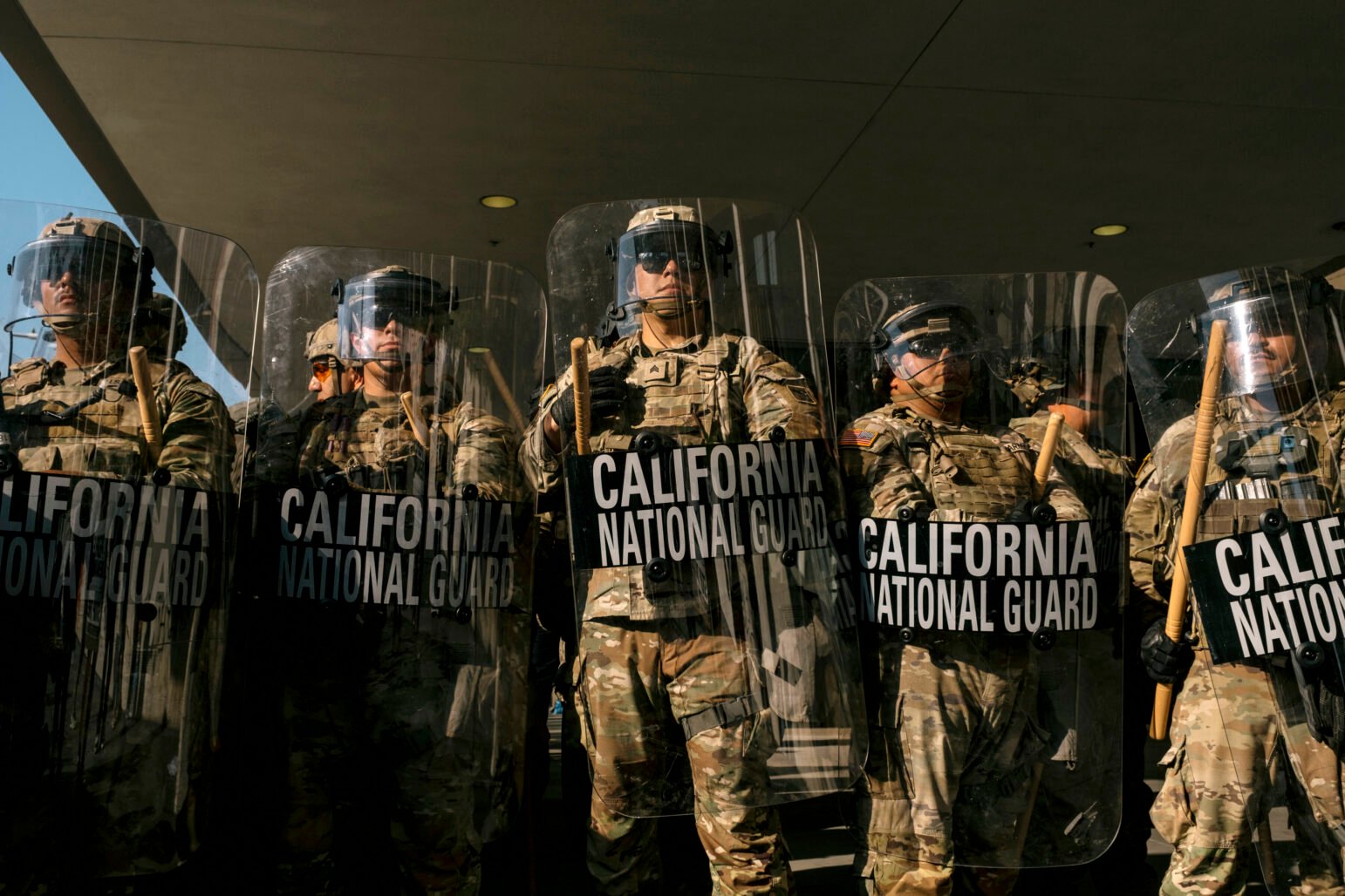 A line of California National Guard members in camouflage uniforms stands shoulder to shoulder holding clear riot shields and wooden batons. The shields are labeled “CALIFORNIA NATIONAL GUARD” in bold white letters. The guardsmen wear helmets, goggles, and protective gear, forming a defensive line under a building overhang.
