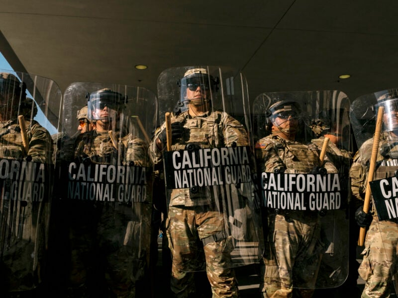 A line of California National Guard members in camouflage uniforms stands shoulder to shoulder holding clear riot shields and wooden batons. The shields are labeled “CALIFORNIA NATIONAL GUARD” in bold white letters. The guardsmen wear helmets, goggles, and protective gear, forming a defensive line under a building overhang.