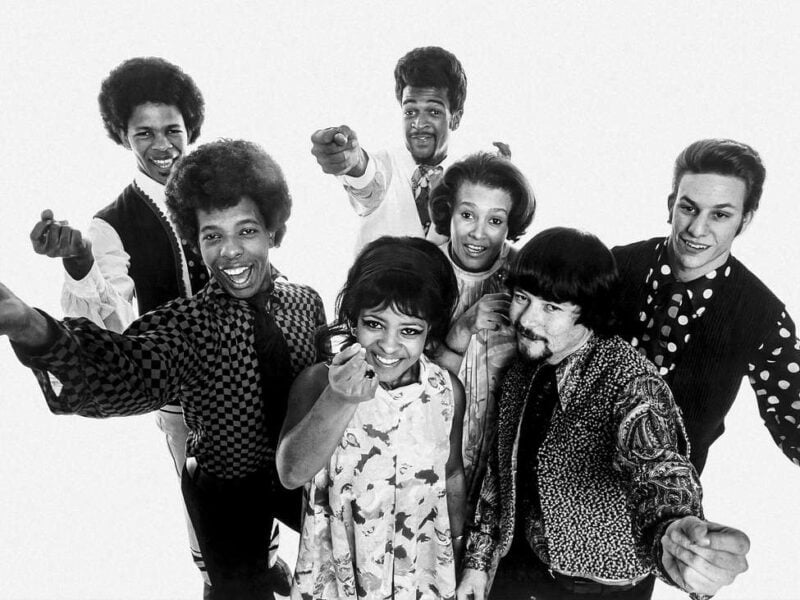 A black-and-white publicity photo from 1968 shows seven members of a band posing enthusiastically toward the camera, smiling and reaching forward. The group wears a mix of patterned and solid outfits typical of the era, radiating a sense of joy and unity against a plain white background.