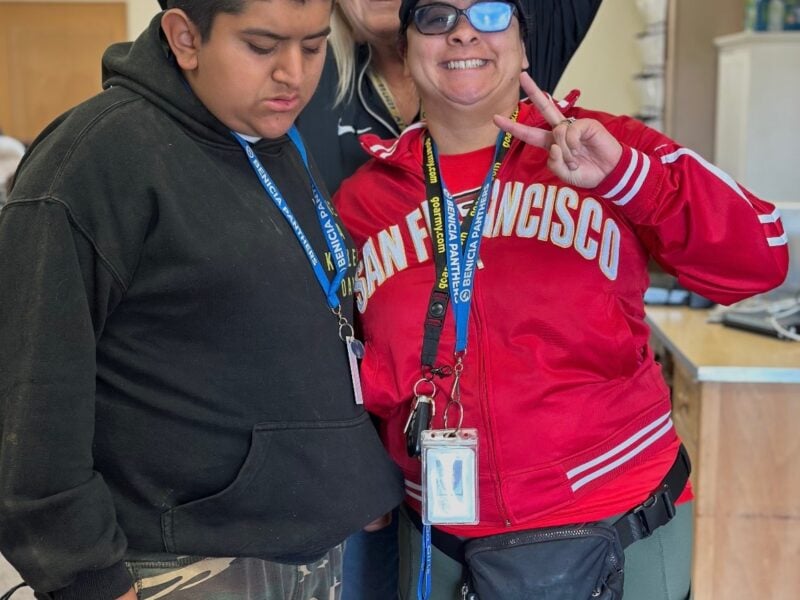 A young boy in camouflage pants and a black hoodie stands beside a woman in a red San Francisco jacket flashing a peace sign. Both wear ID badges. Behind them, a smiling woman points down at the pair from behind.