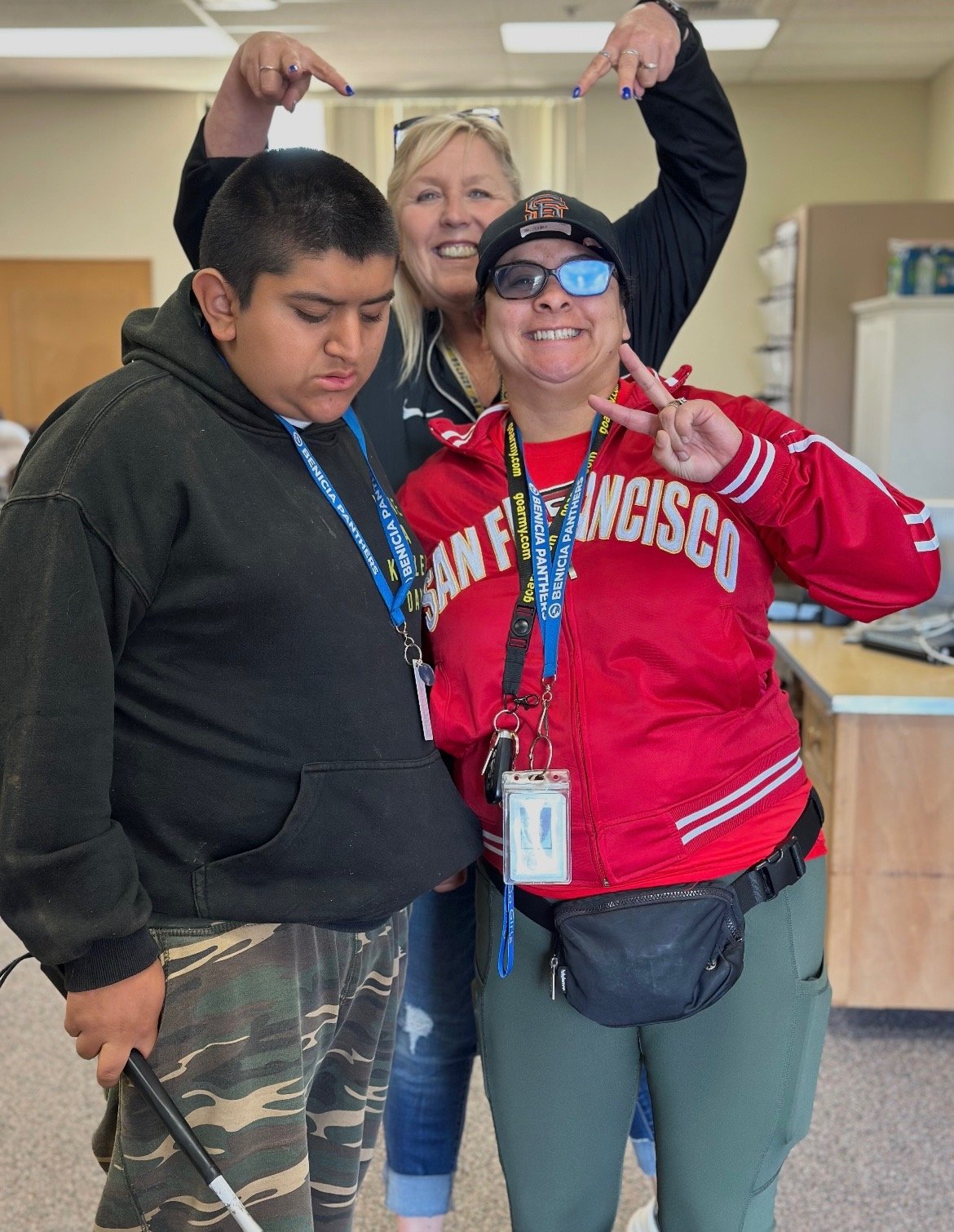 A young boy in camouflage pants and a black hoodie stands beside a woman in a red San Francisco jacket flashing a peace sign. Both wear ID badges. Behind them, a smiling woman points down at the pair from behind.