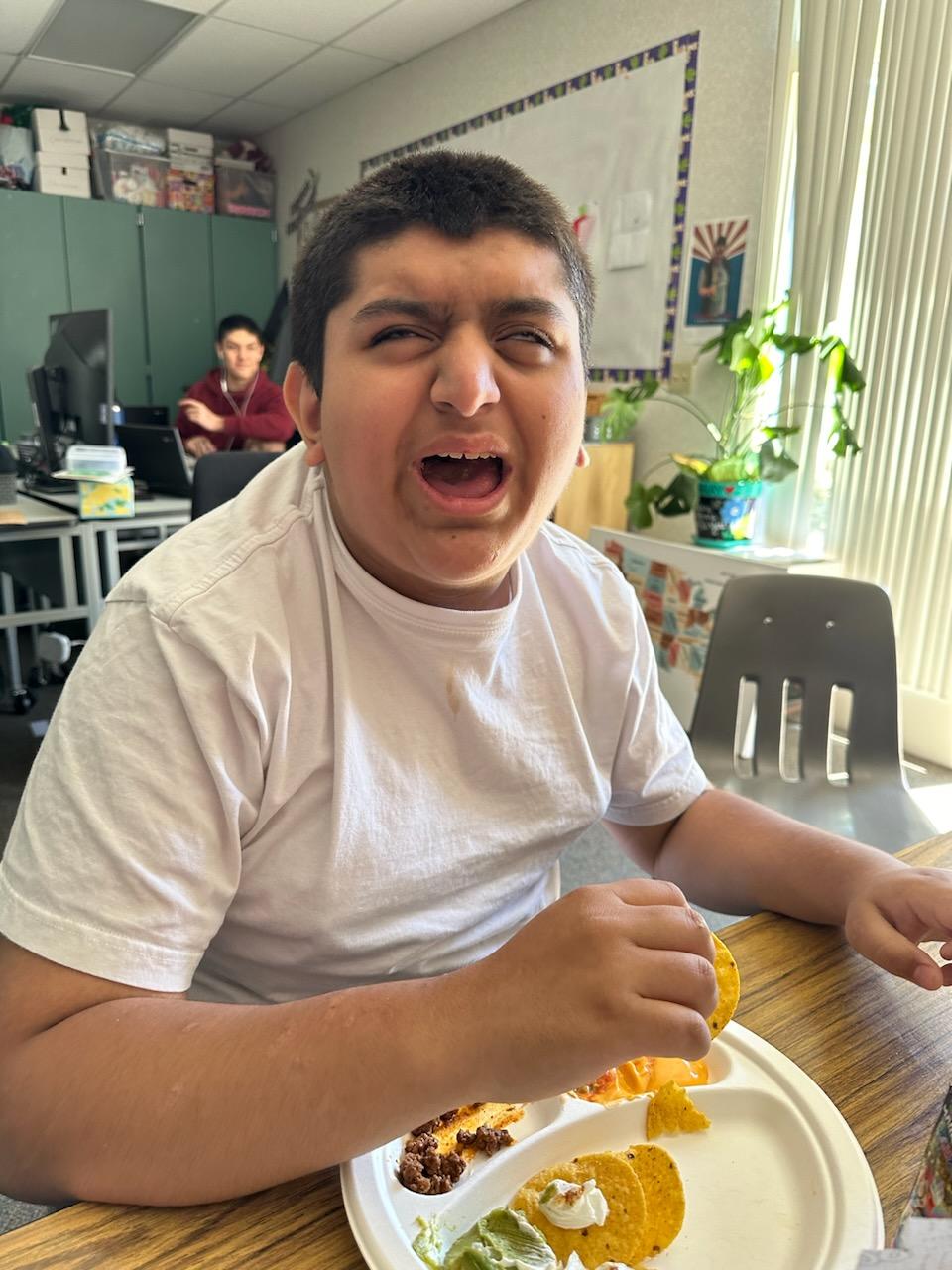 A young boy in a white T-shirt sits at a classroom table eating nachos from a paper plate. He appears to be making a playful face with his mouth open. A classmate is visible in the background.