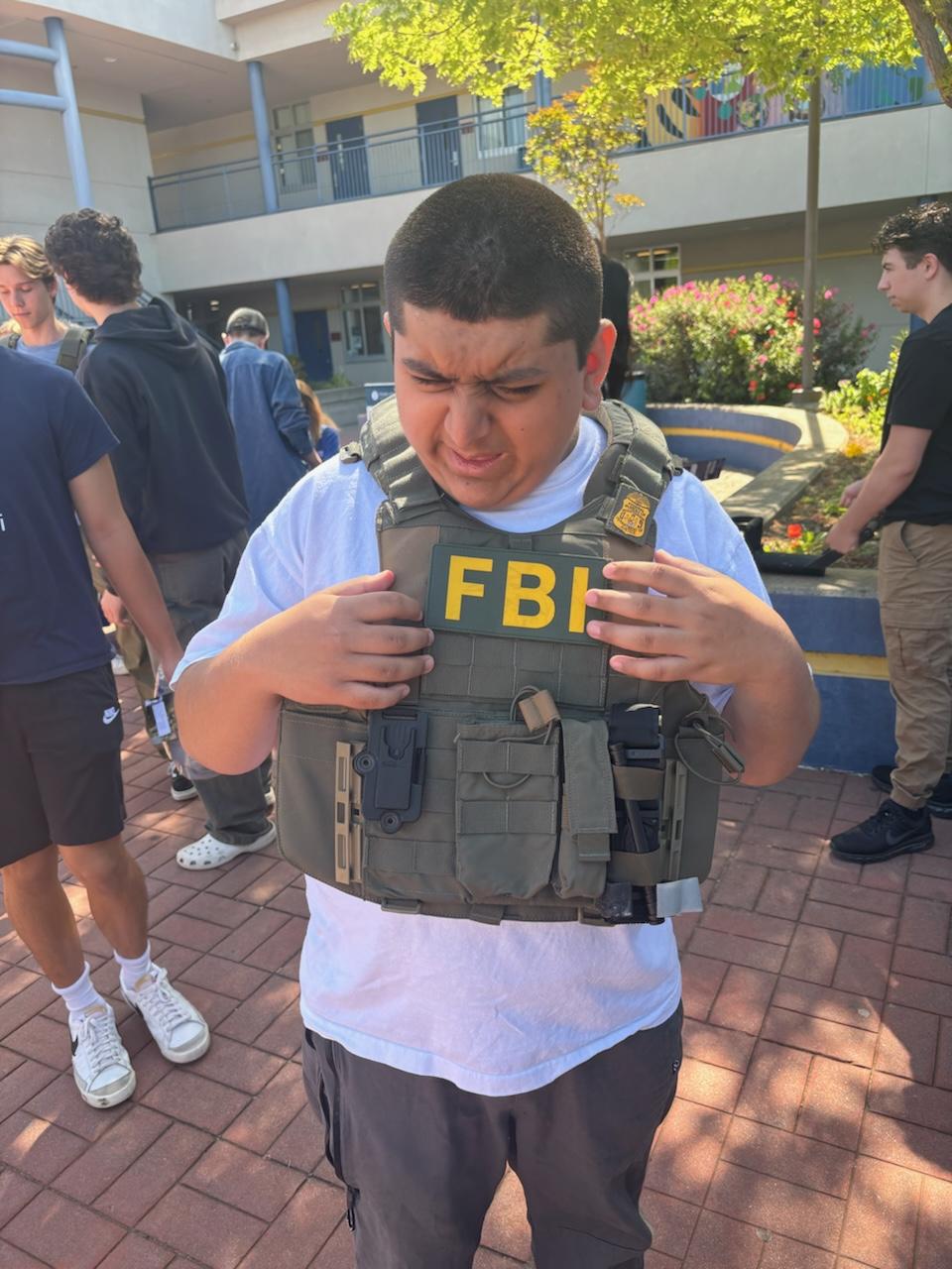 A young boy wears an oversized FBI tactical vest while standing outdoors among a group of students. He squints as he looks down at the vest.