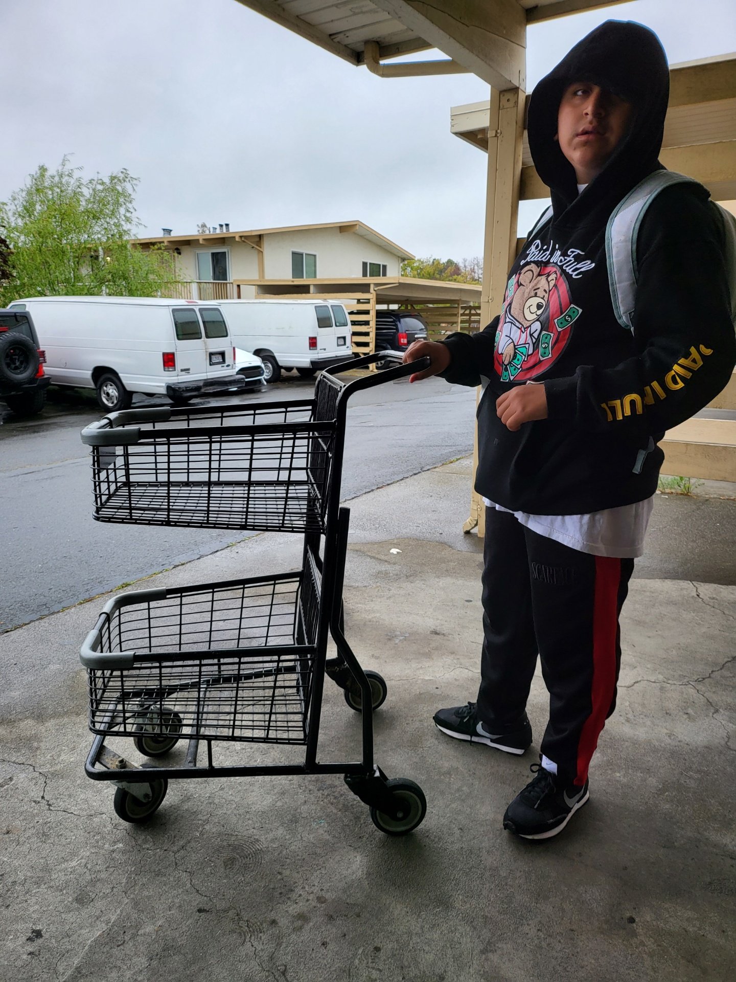 A young boy wearing a hoodie, sweatpants, and a backpack stands outside with a two-tier rolling cart. Several white vans and apartment buildings are in the background.