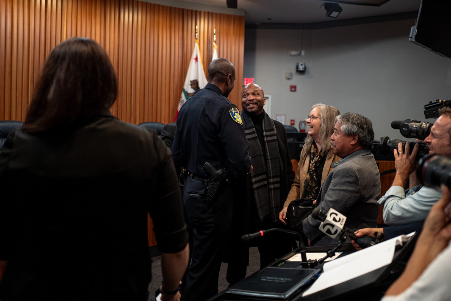 A uniformed police chief warmly interacts with smiling community members in a government chamber, as photographers and media personnel capture the moment. The background shows flags, a wood-paneled wall, and official meeting desks, indicating a civic or public event.