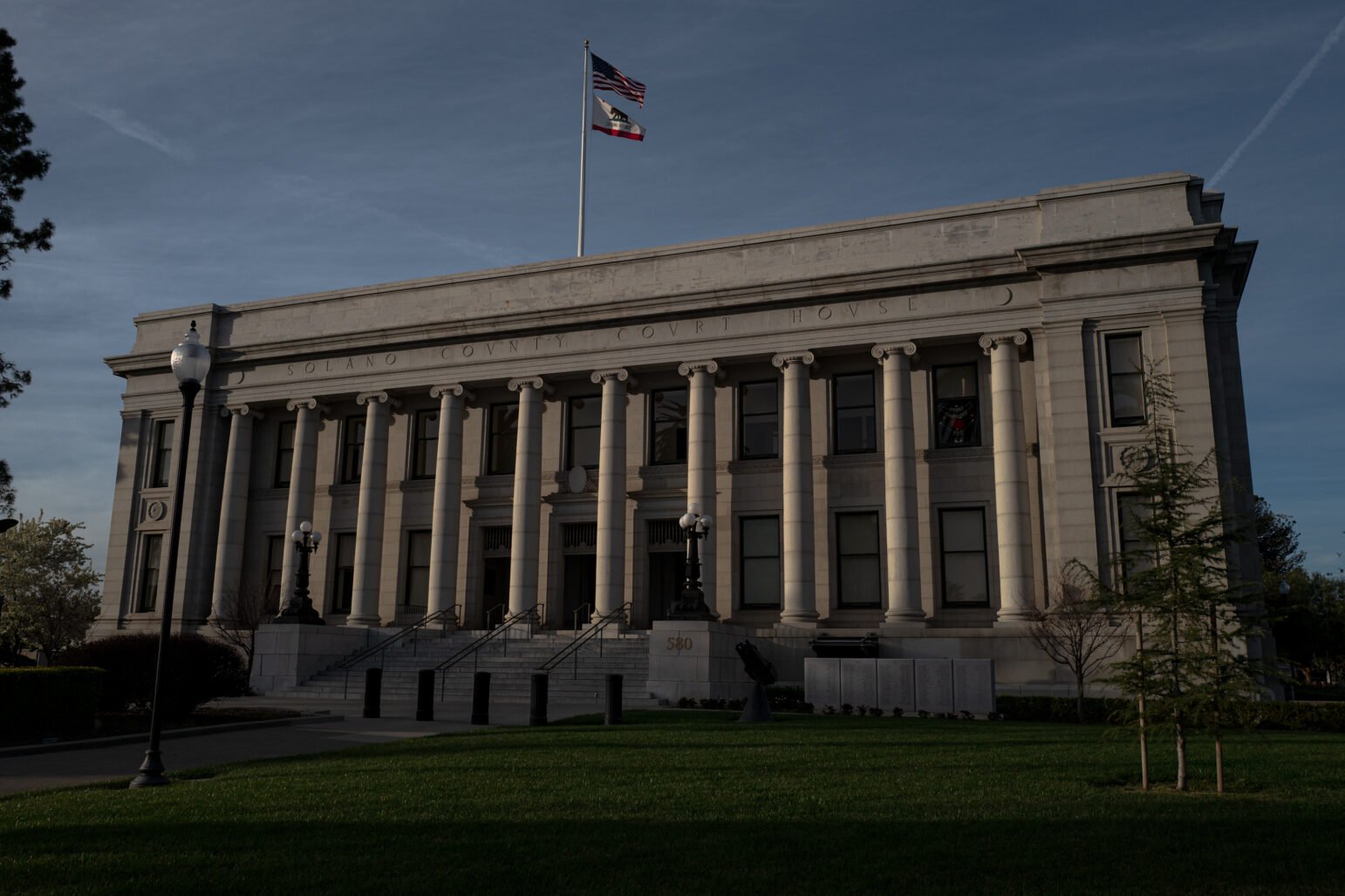 The Solano County Courthouse in Fairfield, California, stands under a clear sky with American and California state flags flying above its neoclassical facade, flanked by tall columns and a manicured lawn in the foreground.