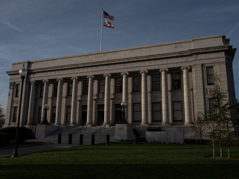 The Solano County Courthouse in Fairfield, California, stands under a clear sky with American and California state flags flying above its neoclassical facade, flanked by tall columns and a manicured lawn in the foreground.