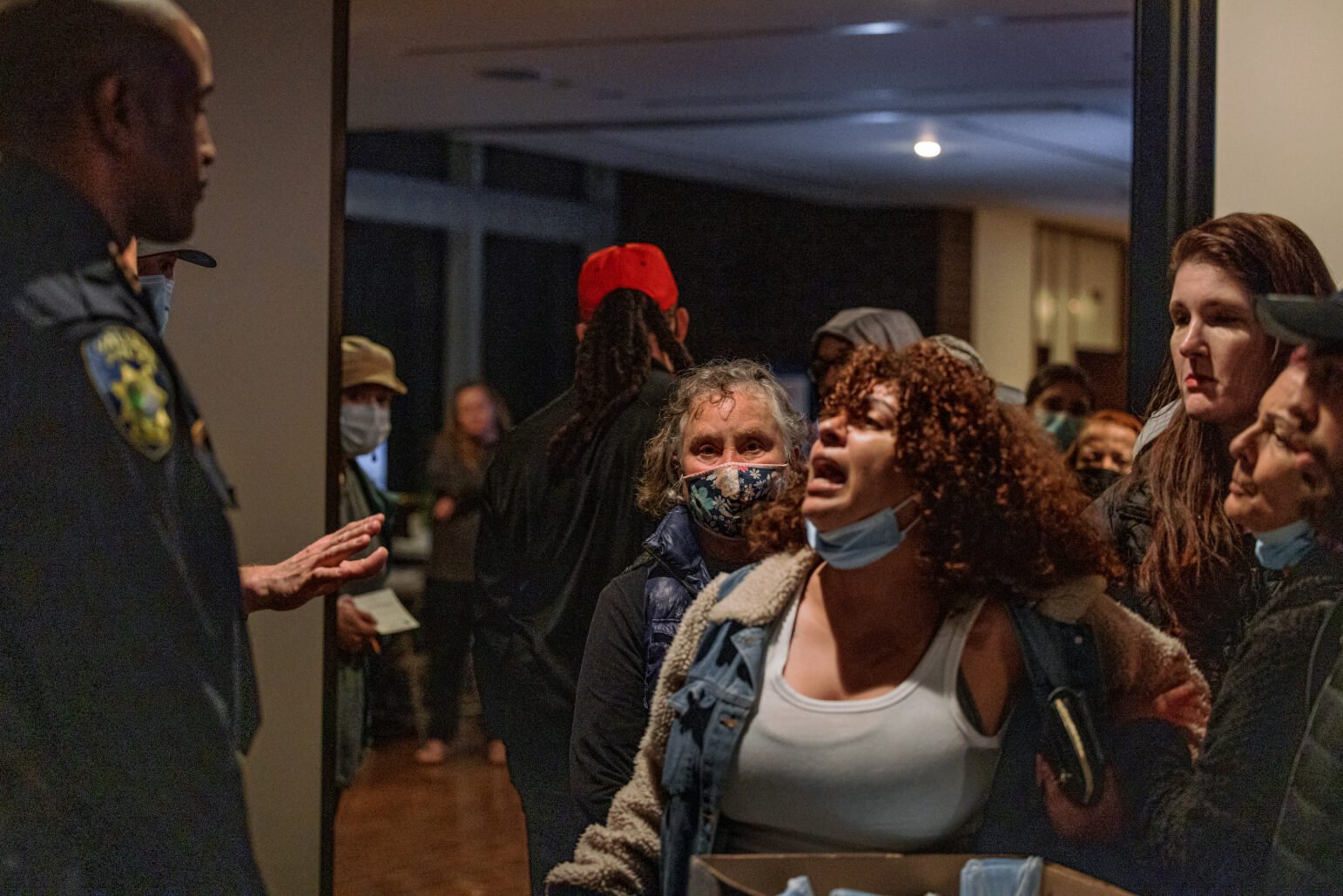 A group of community members confront a uniformed Vallejo police officer inside City Hall. One woman shouts emotionally while others crowd the doorway behind her, some wearing masks and others visibly distraught.
