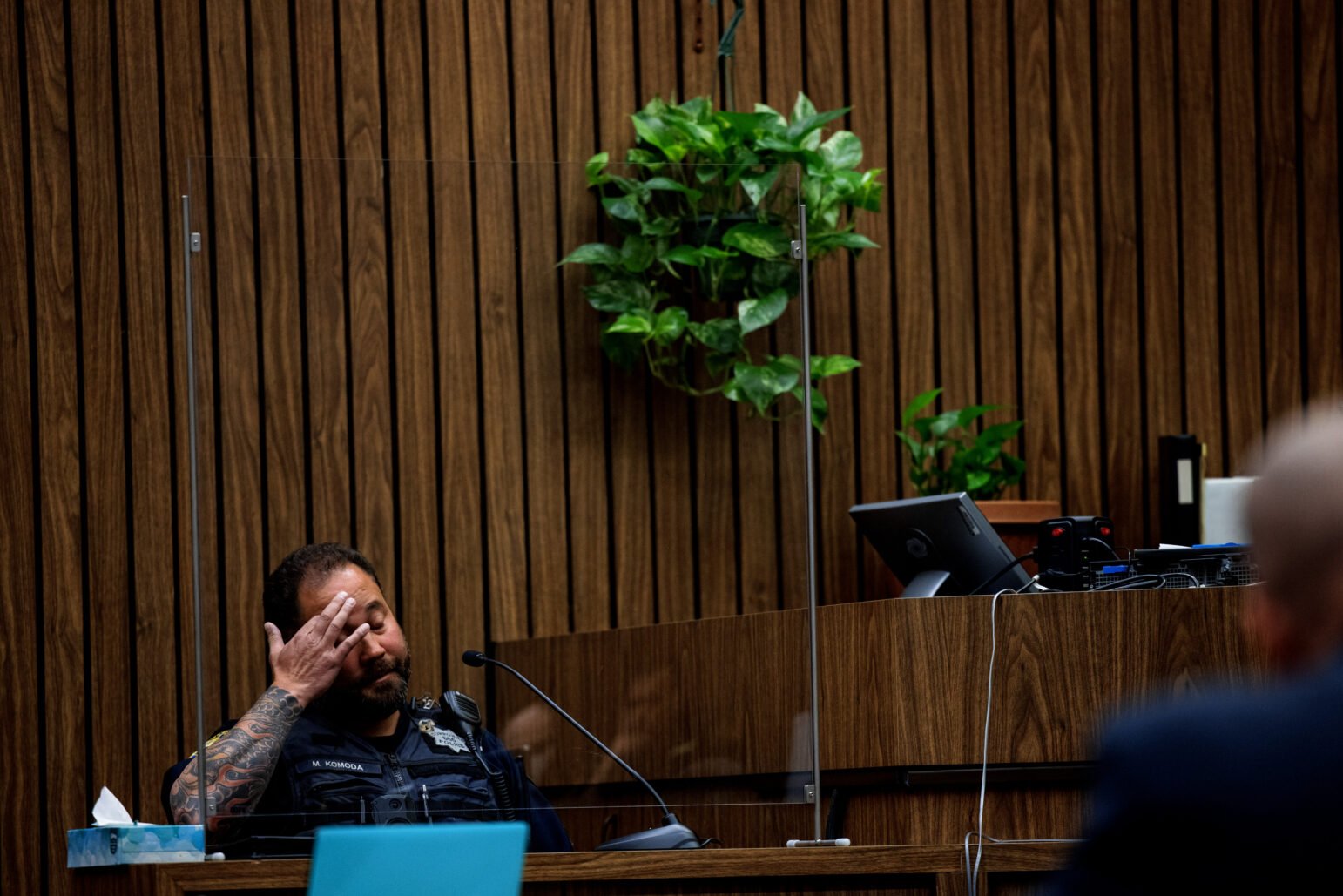 A uniformed police officer with visible arm tattoos sits behind a transparent barrier on a courtroom witness stand. The wood-paneled wall behind him features hanging green plants, and courtroom equipment is visible nearby.